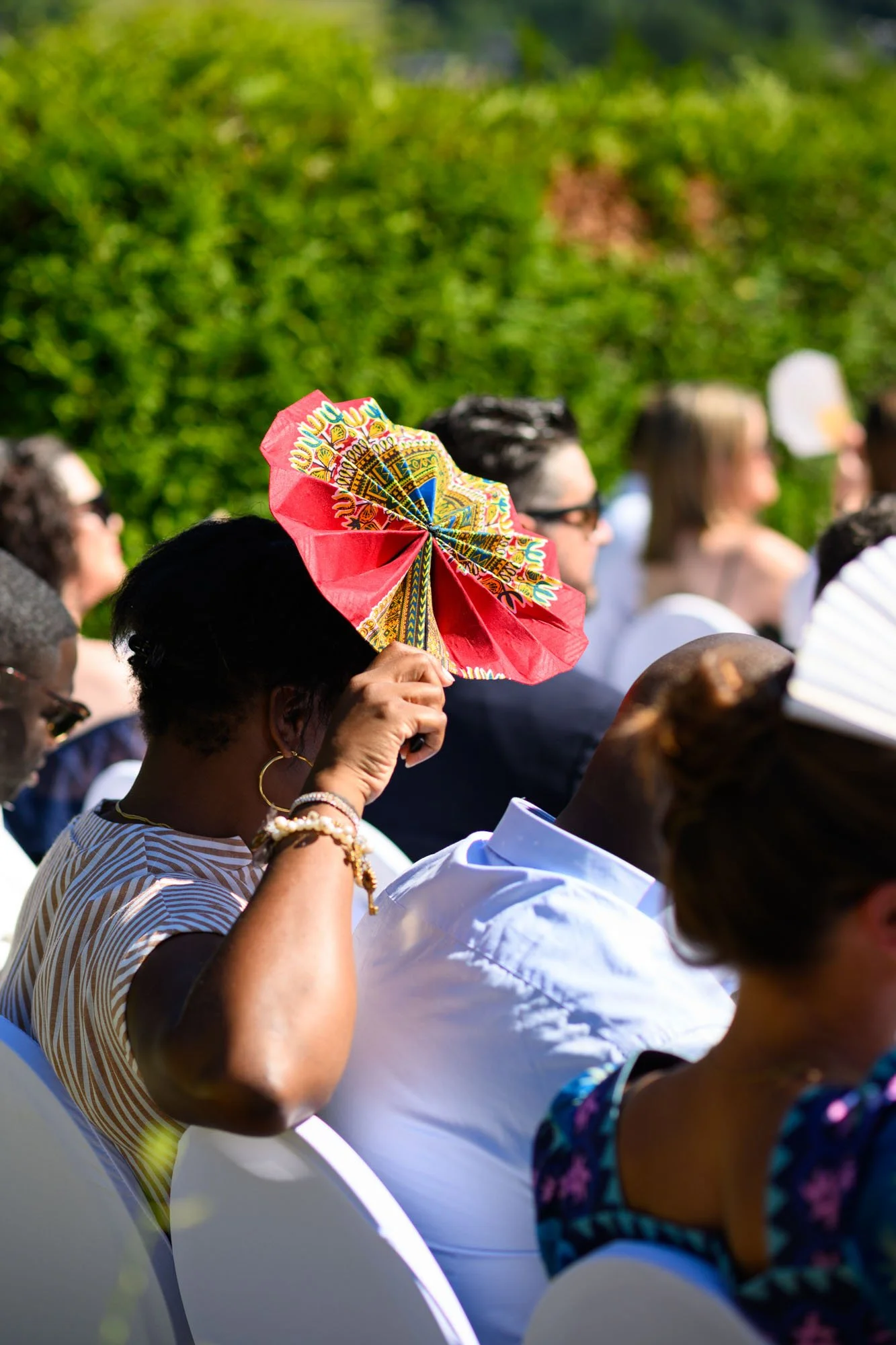 Foule de personnes assises en extérieur lors d'un événement, avec une femme en premier plan tenant un éventail coloré en forme de fleur devant son visage, derrière un écrin de verdure.