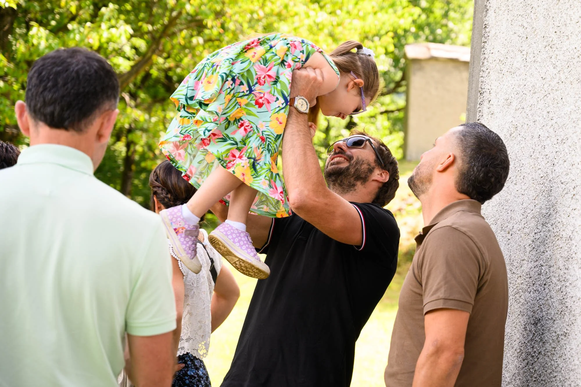 Un homme souriant tient une jeune fille dans ses bras lors d'un rassemblement en plein air avec plusieurs autres personnes, dont deux hommes, devant un mur et entourés de verdure.