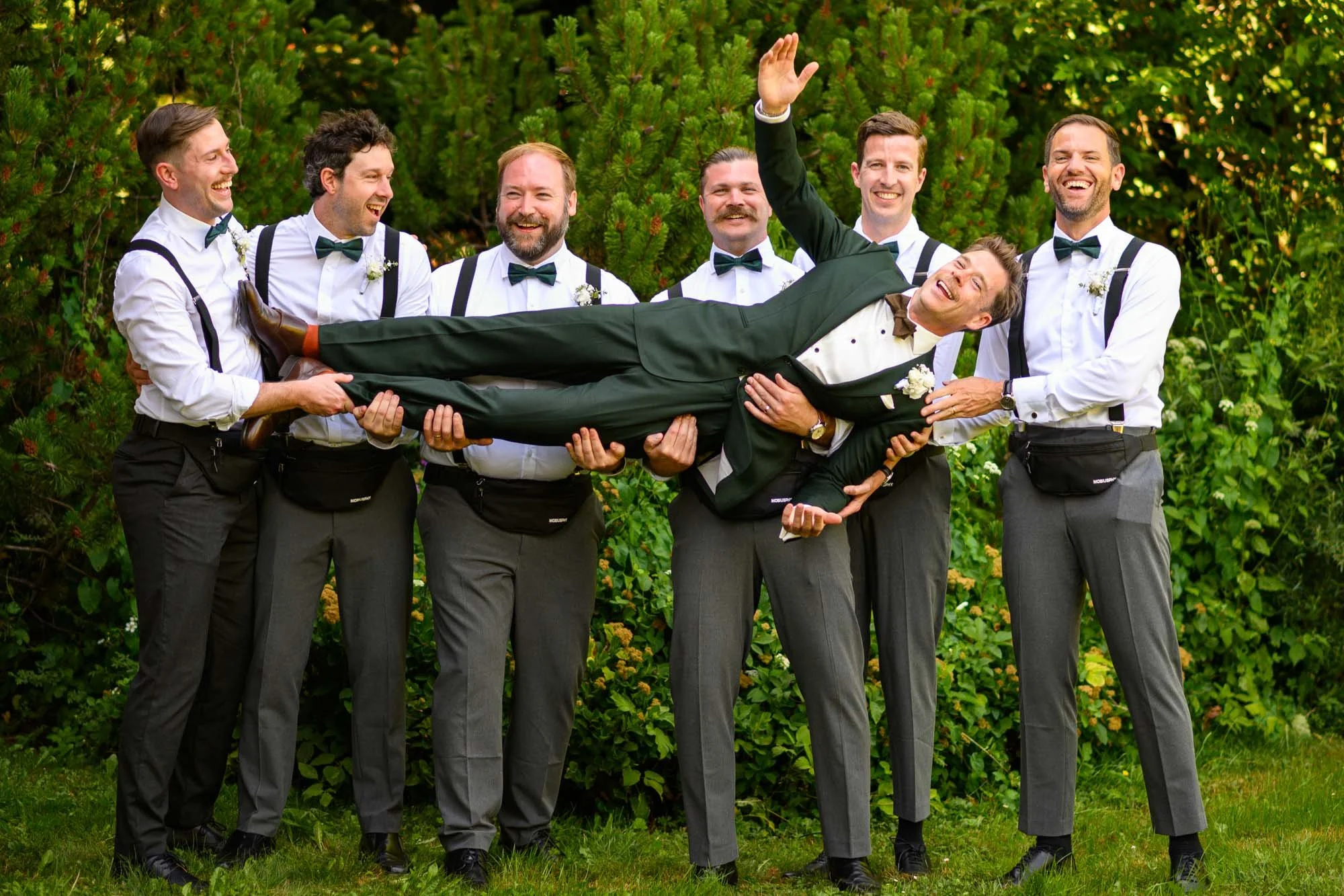 Un groupe d'hommes en costume tenant un homme en costume de mariage horizontalement, tous rient en arrière-plan un buisson vert luxuriant.