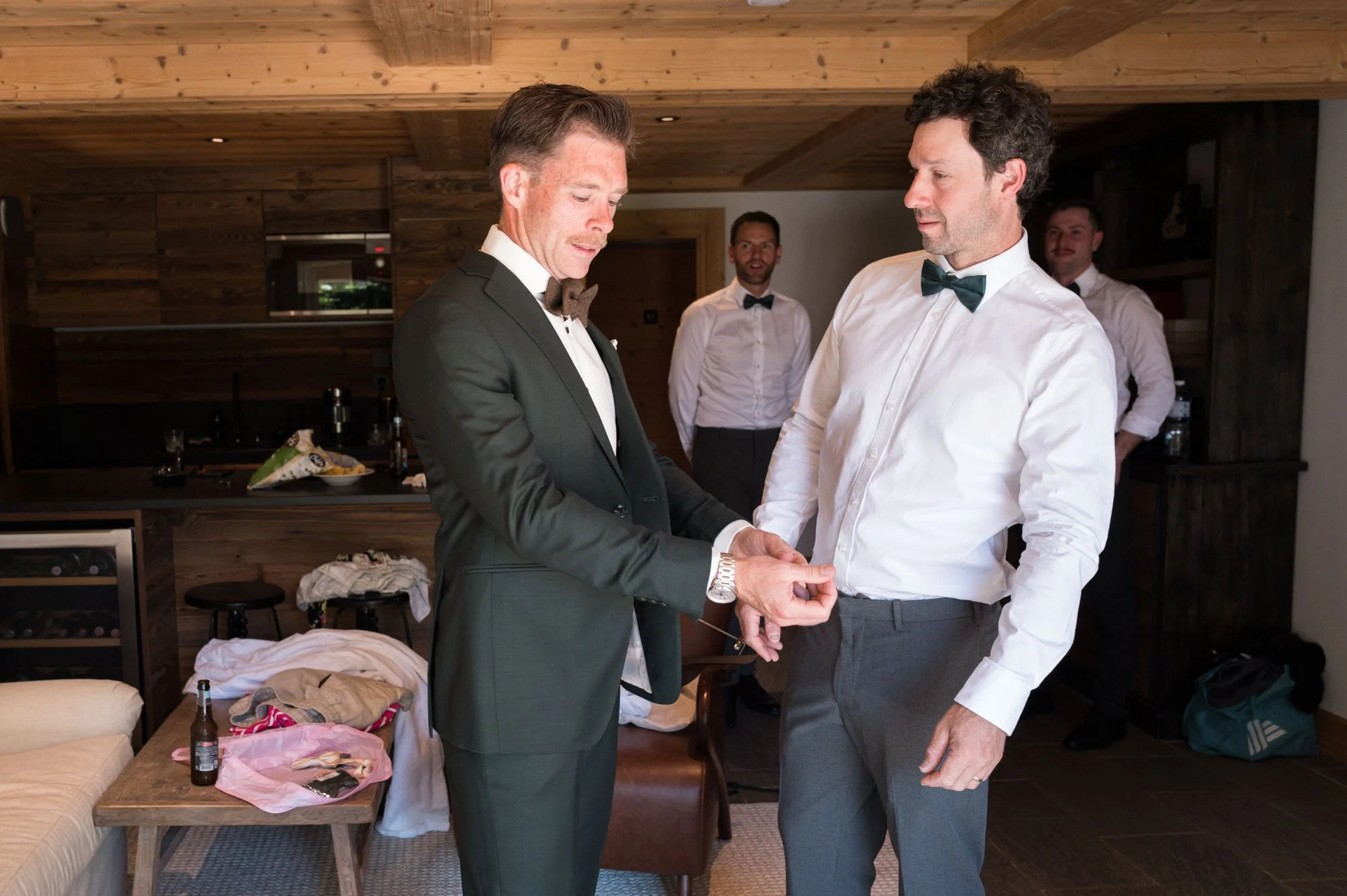 Deux hommes habillés en costumes de mariage, en train de se joindre la main dans une pièce en bois avec trois autres hommes en arrière-plan