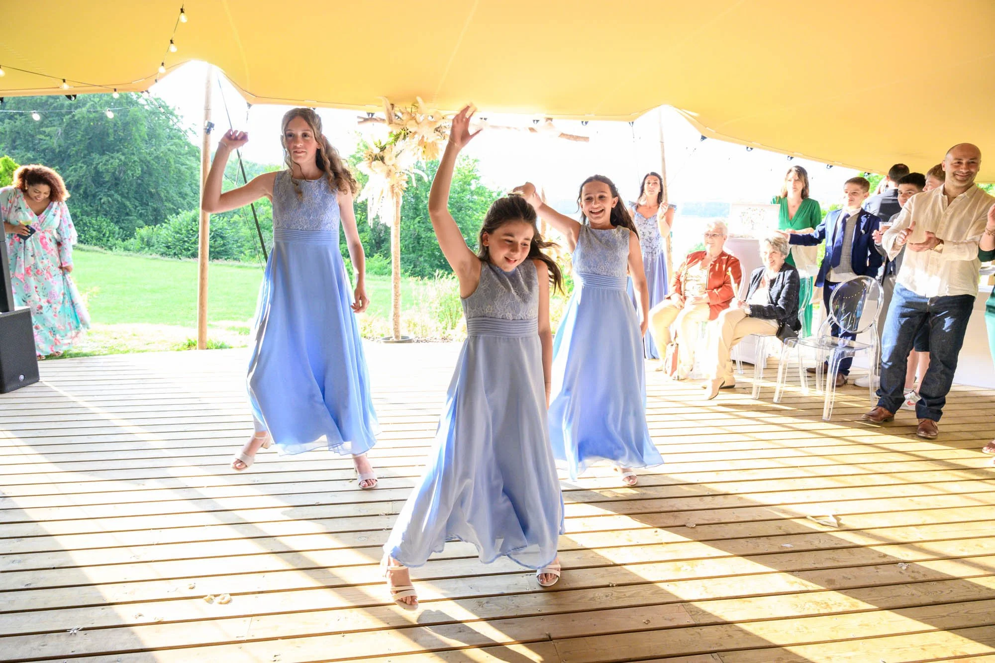 Jeunes filles dansant et souriant sous une tente lors d'une fête en plein air, avec des invités assis en arrière-plan.