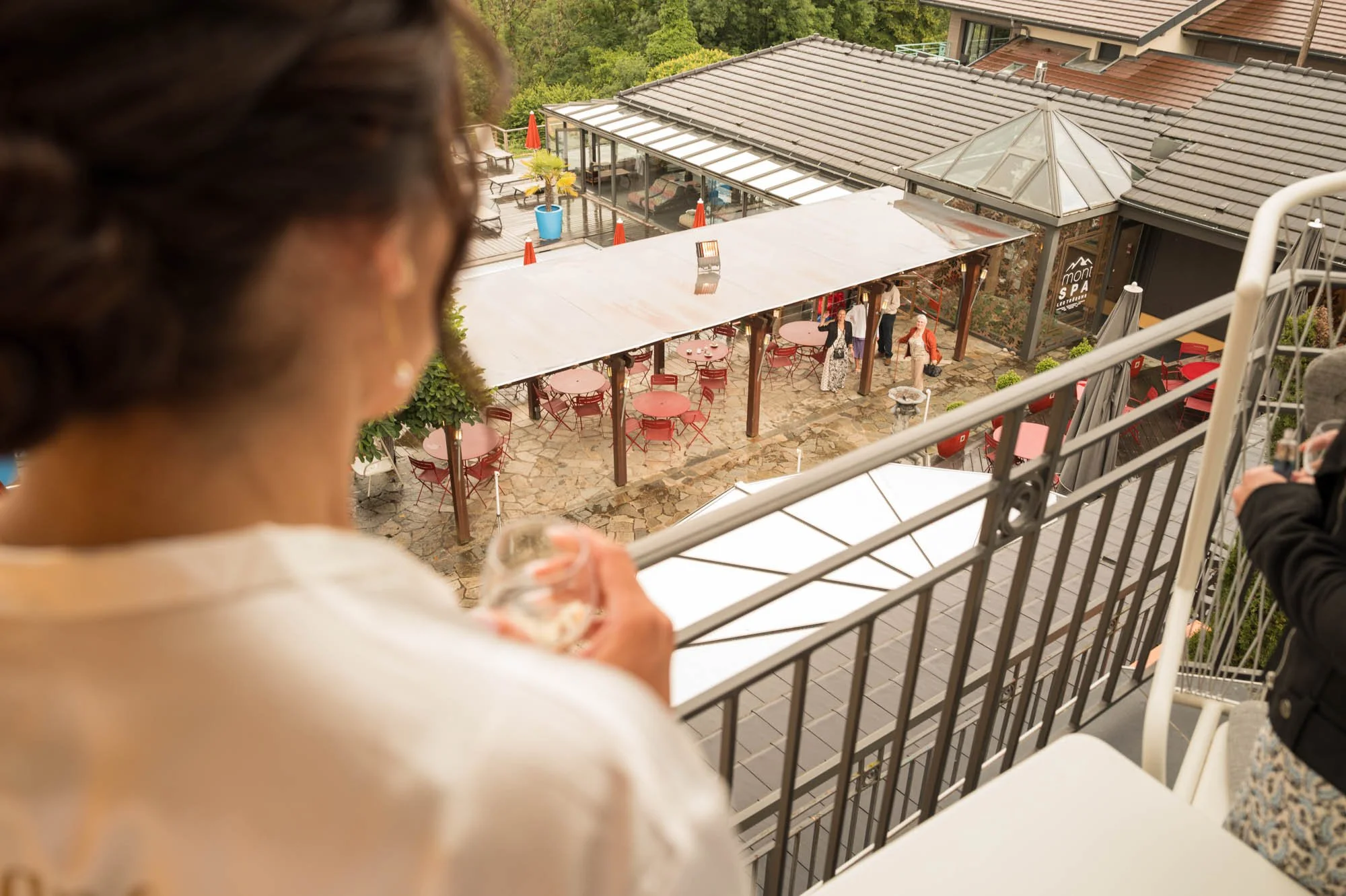 Vue d'en haut d'une terrasse de restaurant avec des tables rondes et des chaises, des gens marchant et une structure en verre, avec un bâtiment à toiture en tuiles en arrière-plan.