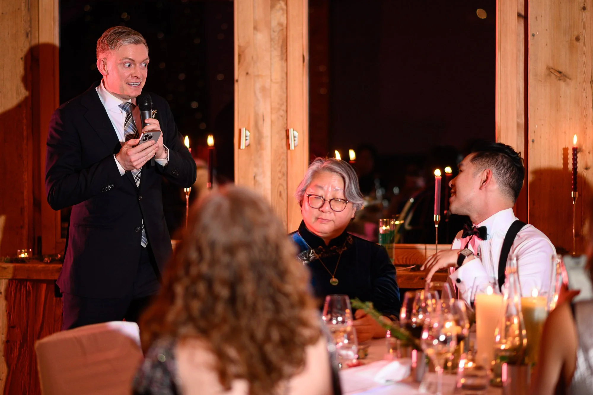Un homme en costume noir parle à un microphone lors d'une réunion ou d'un événement dans une salle avec des personnes assises autour d'une table, dans une ambiance chaleureuse et festive.
