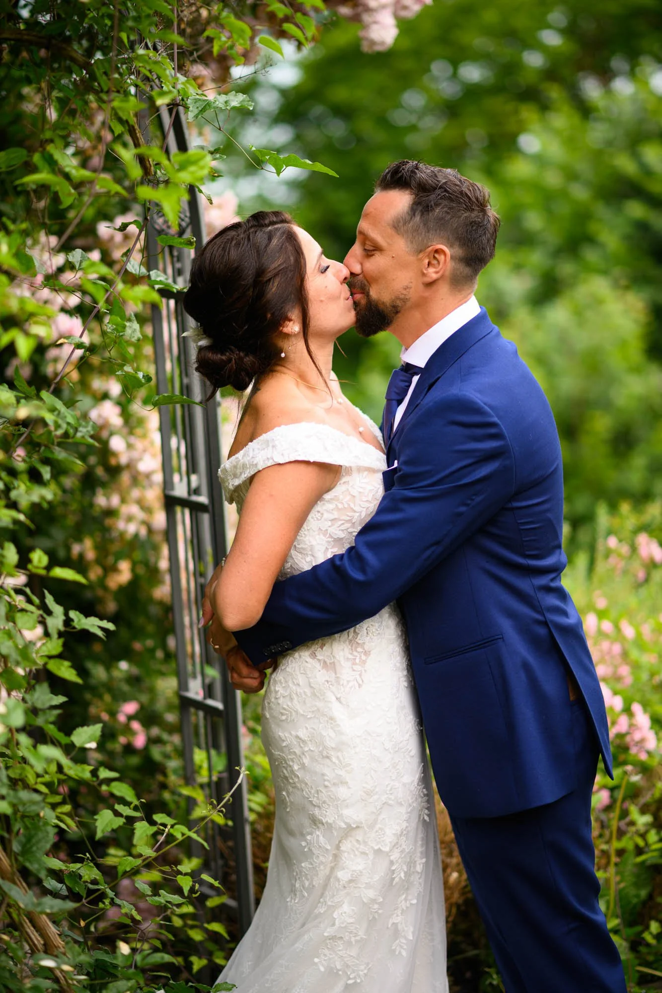Un couple de mariés s'embrassant dans un jardin verdoyant, avec des fleurs roses en arrière-plan.