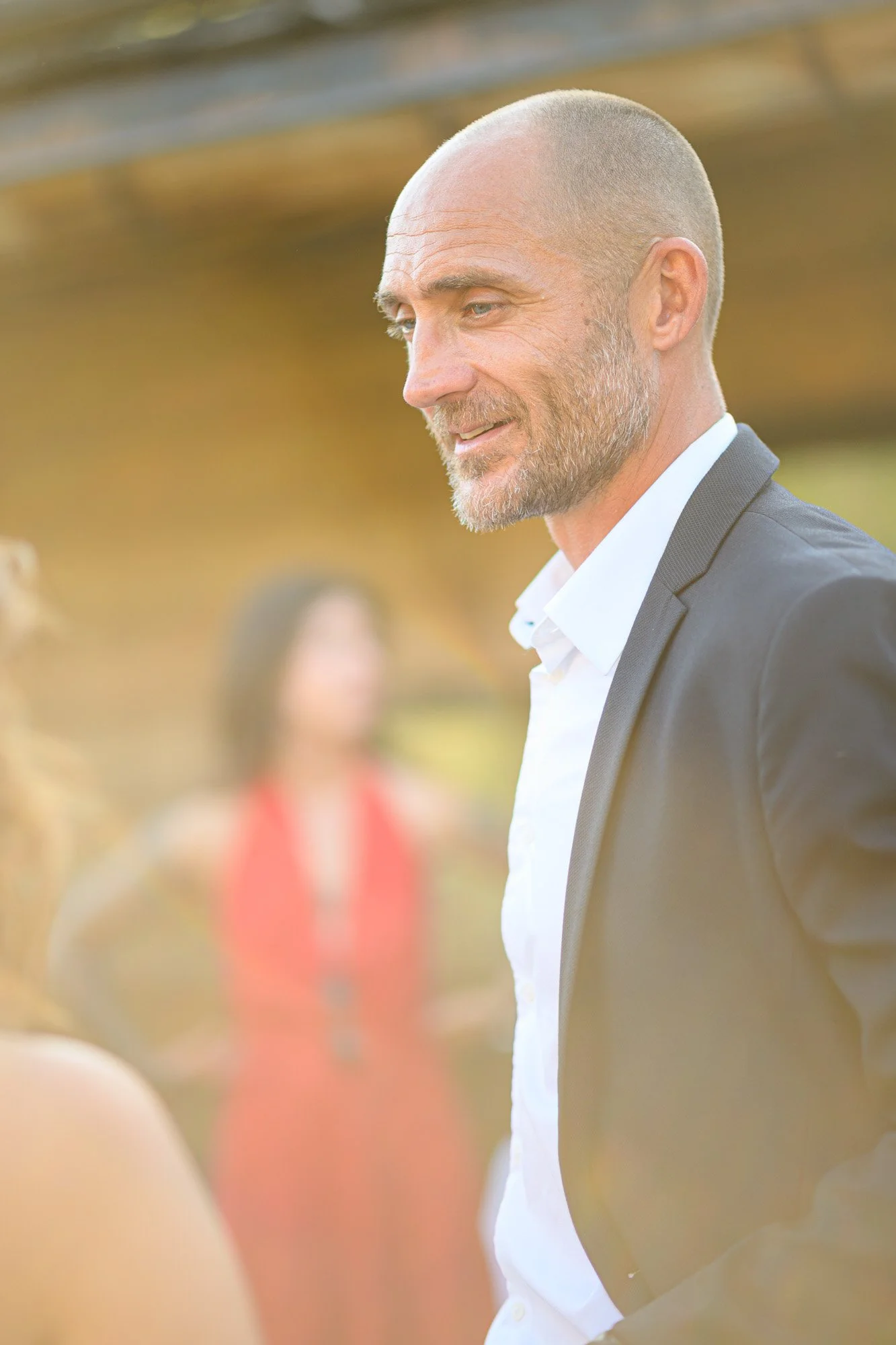 Un homme souriant et en costume, lors d'un événement en plein air avec des personnes en arrière-plan.