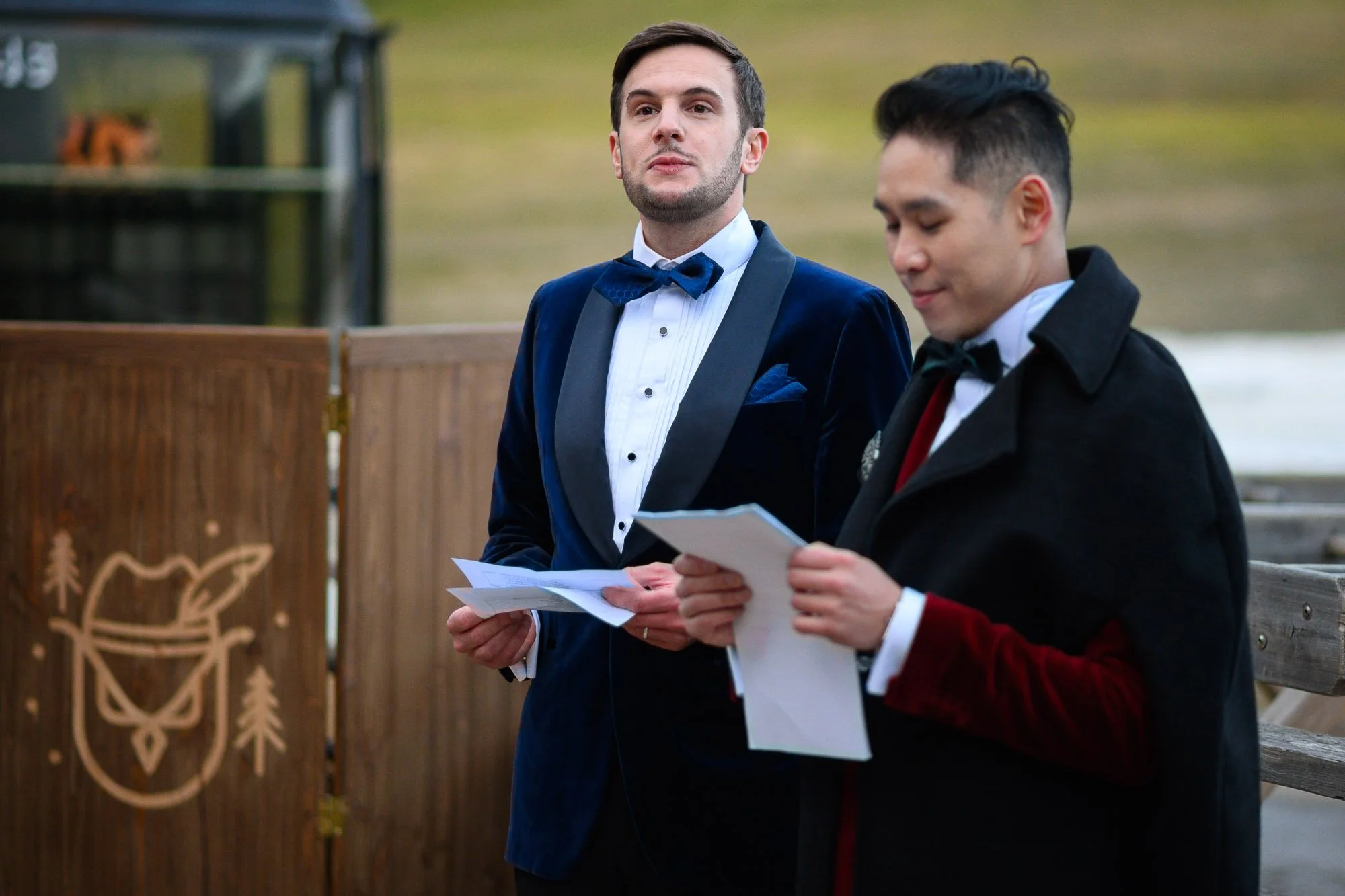 Deux hommes portant des costumes élégants, lisant des documents, se tiennent à l'extérieur près d'une barrière en bois.