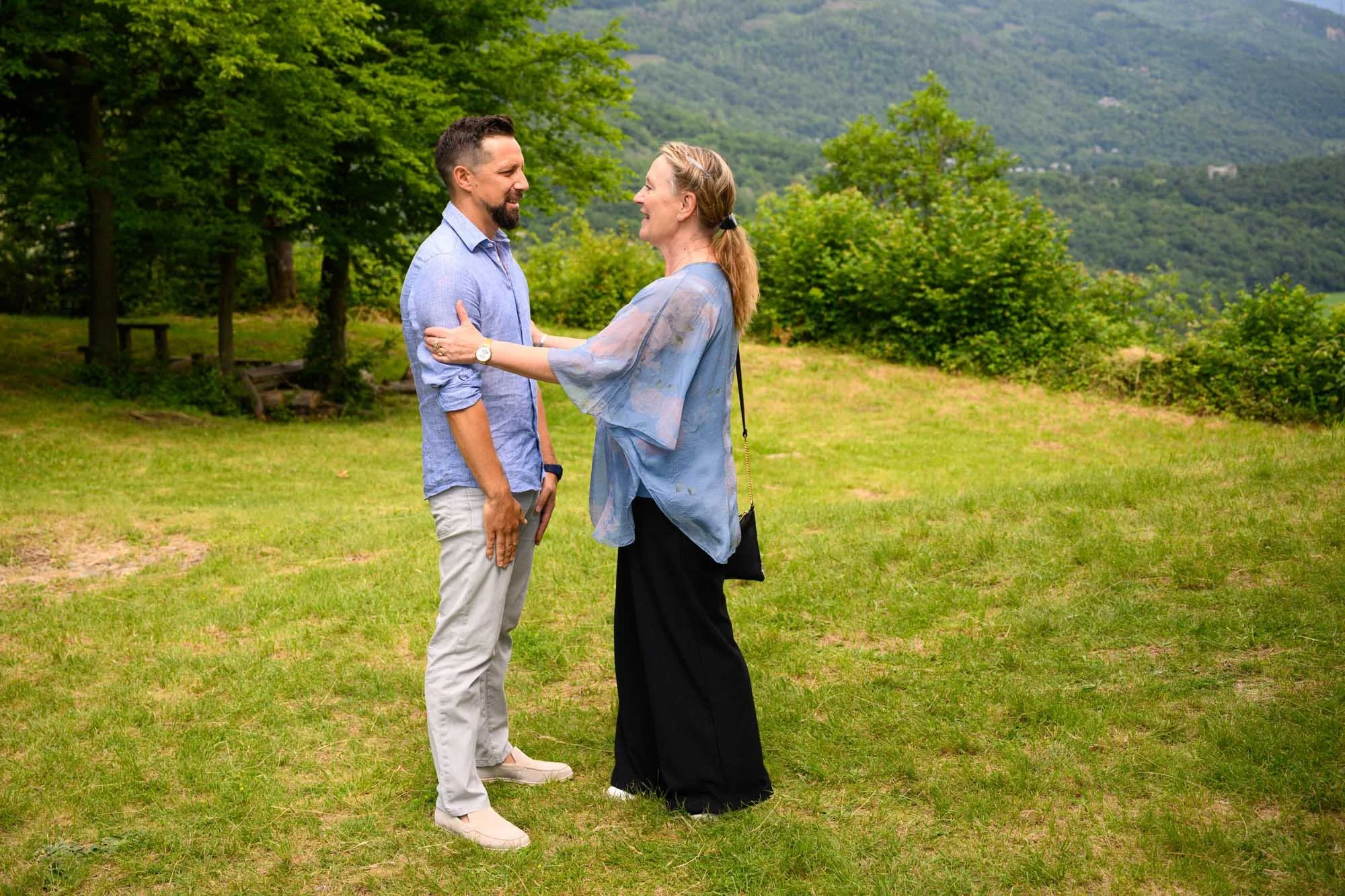 Un couple se tient face à face dans un champ vert, souriant et se regardant dans les yeux, avec un paysage de montagnes en arrière-plan.