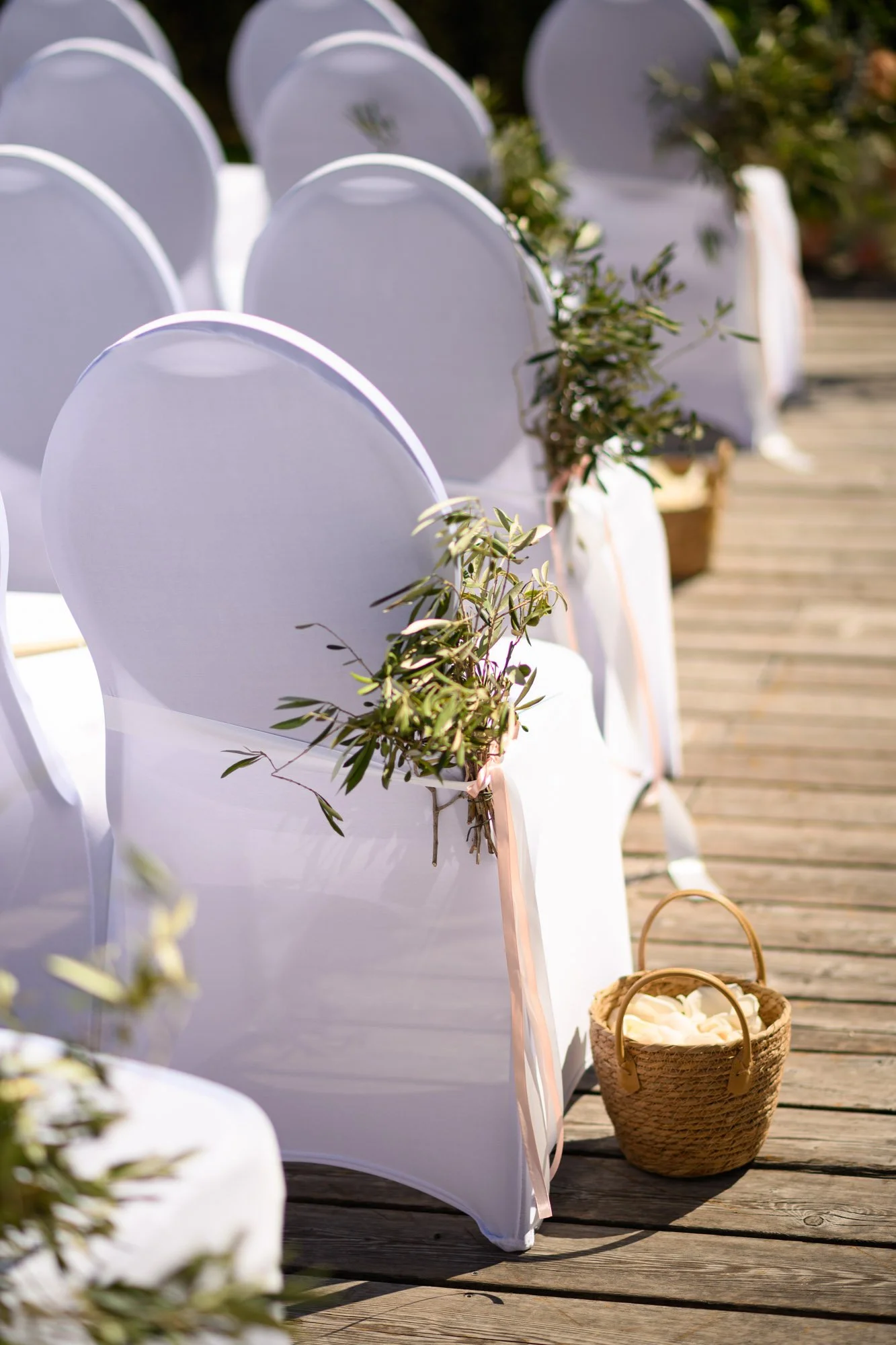 Chaise décorée pour un mariage avec un voile blanc et une branche d'olivier attachée, posée sur une terrasse en bois avec un panier en osier à côté.