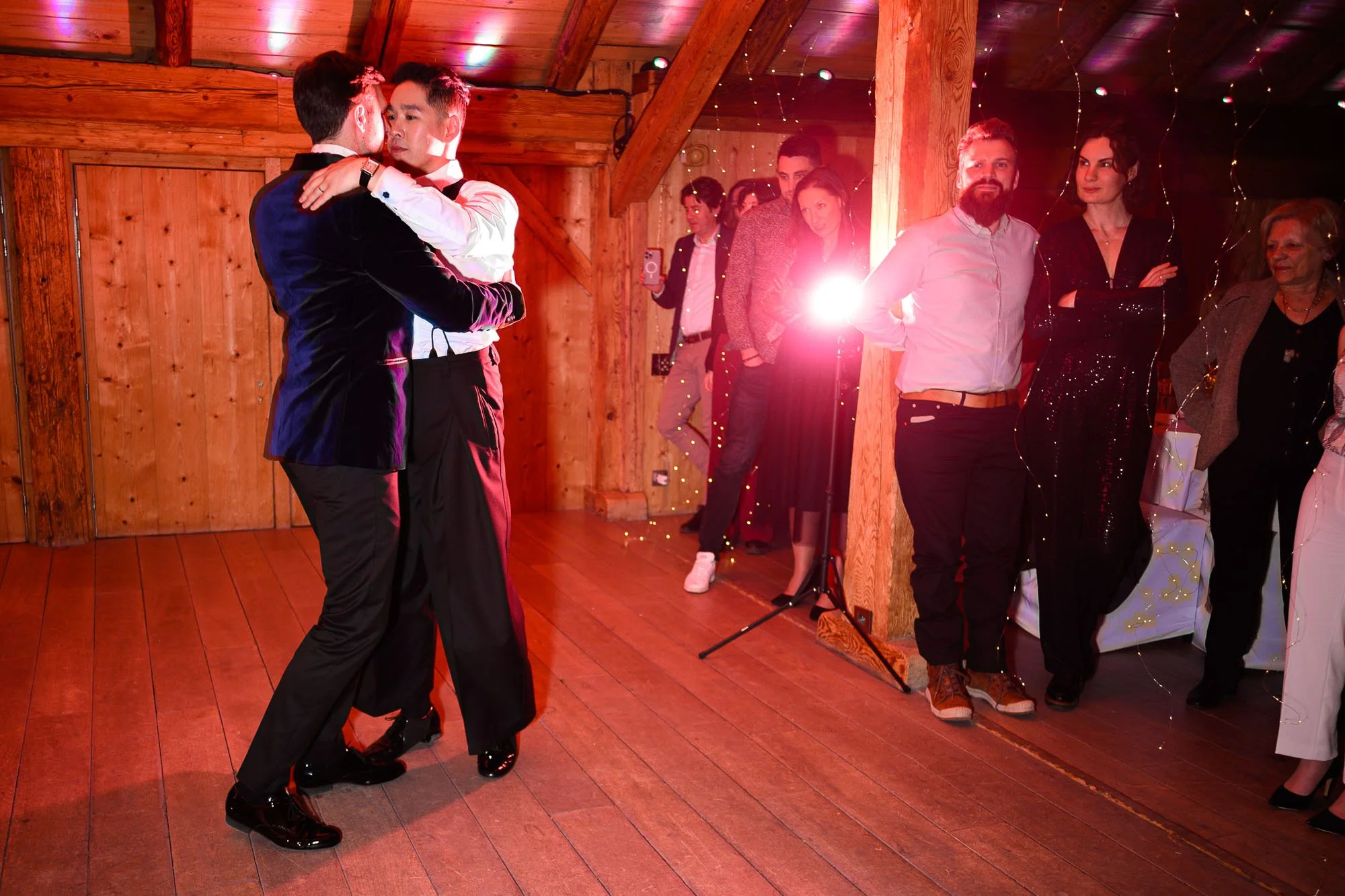 Deux personnes dansent lors d'une fête ou d'un mariage dans une salle en bois. D'autres invités observant la danse se tiennent au fond, certains prenant des photos ou vidéos. L'atmosphère est chaleureuse et festive.
