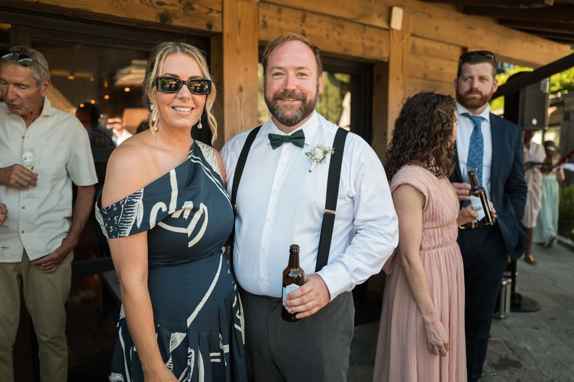 Groupe de personnes lors d'une célébration en extérieur, dont un homme avec nœud papillon et un homme avec bretelles, tous deux tenant une bouteille.