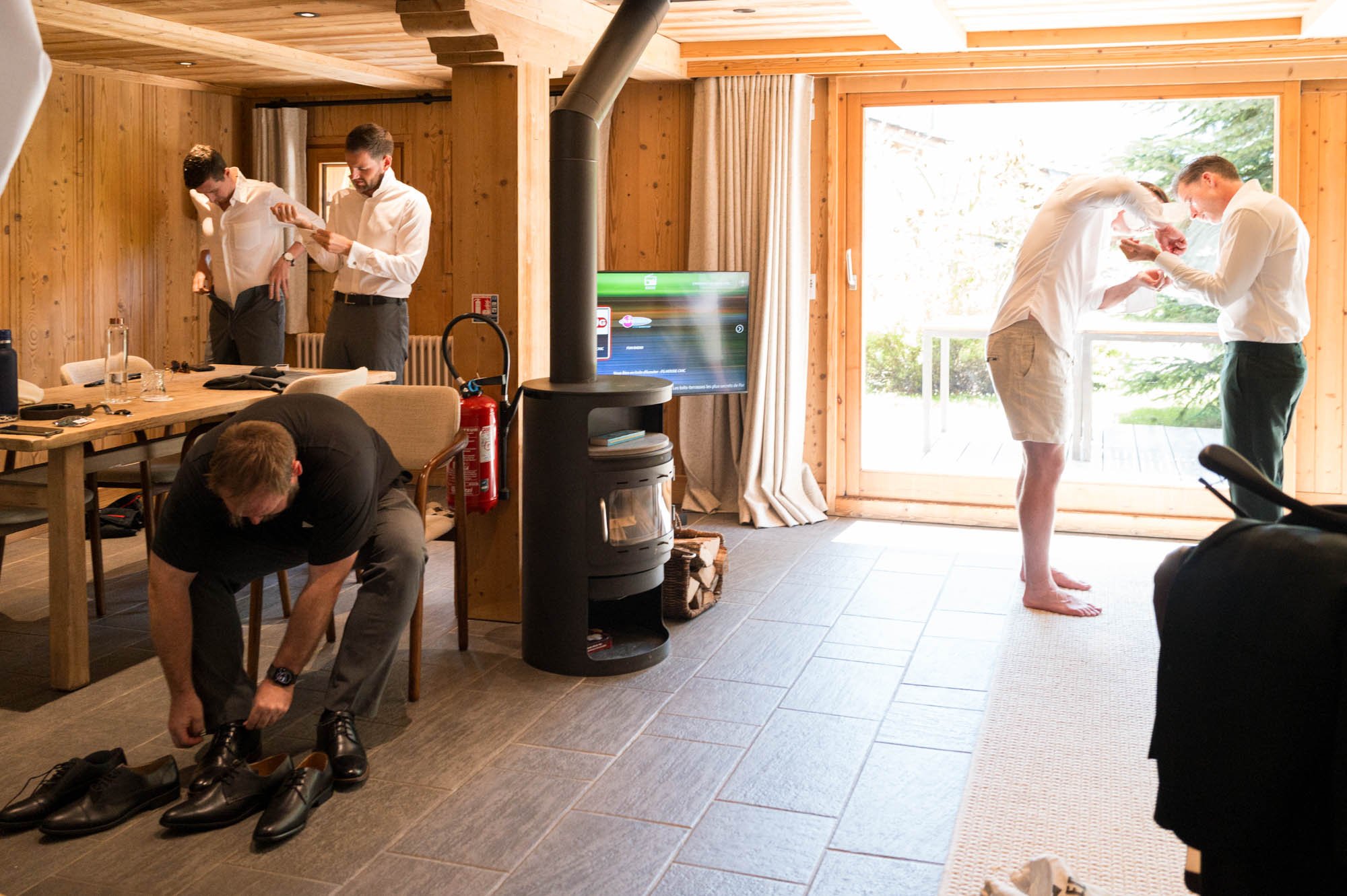 Groom and best man examining a ring, others putting on shoes, and a person bending down to tie shoelaces in a cozy wooden room with large windows and outdoor scenery.