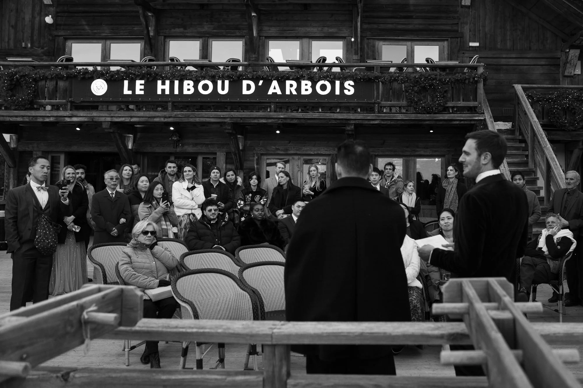 Une scène d'événement en plein air avec un groupe de personnes assises ou debout, écoutant deux hommes en costume qui semblent faire une présentation ou un discours, devant un bâtiment en bois avec un panneau indiquant 'LE HIBOU D'ARBOIS'.