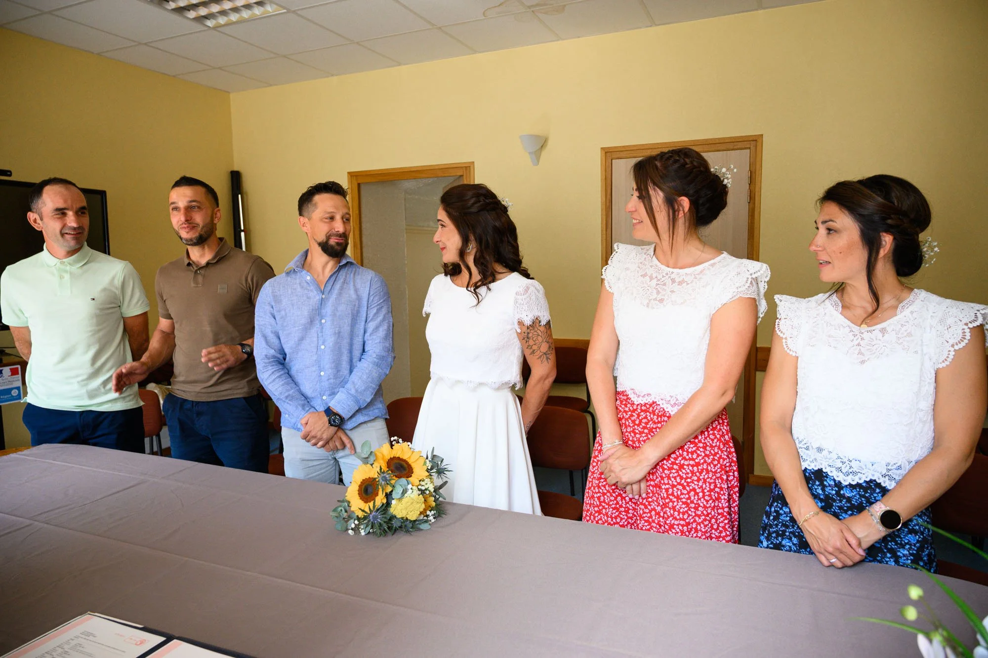 Groupe de personnes lors d'une cérémonie, avec une femme au centre tenant un bouquet de fleurs, entourée d'hommes et de femmes souriants, dans une salle lumineuse.