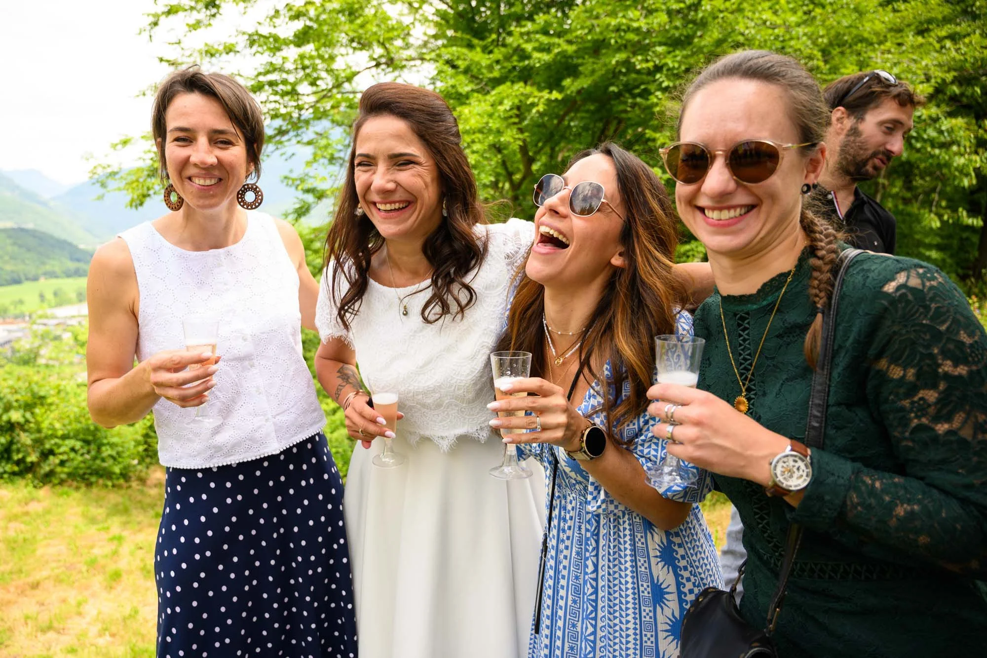 Groupe de cinq personnes souriantes, portant des lunettes de soleil, se tenant par l'épaule, tenant des verres à boisson, dans un paysage verdoyant en plein air.