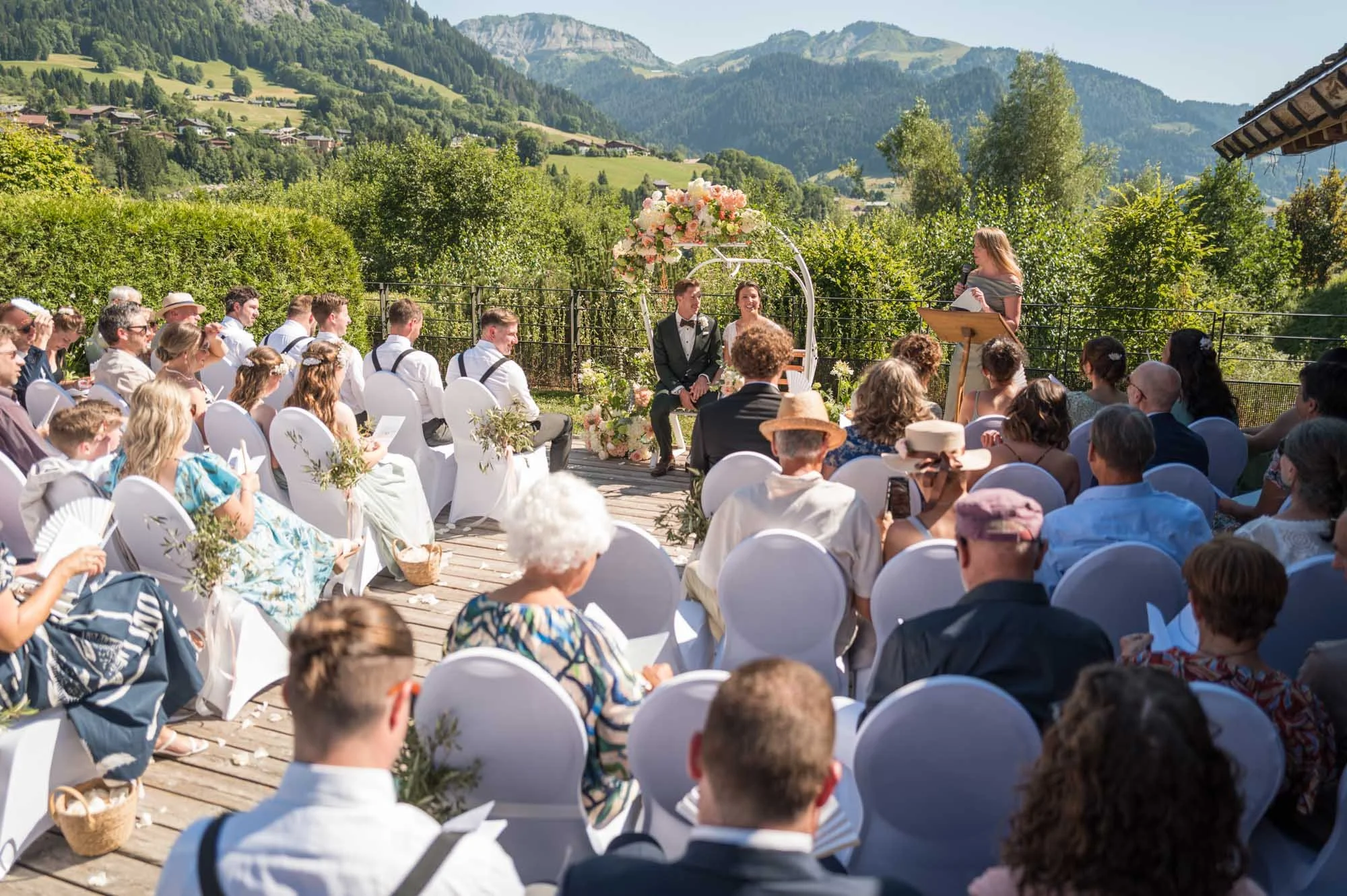 Un mariage en plein air dans un environnement montagneux avec des invités assis, un couple de mariés sous une arche fleurie, une officiante parlant, et un décor floral visible avec un ciel ensoleillé.