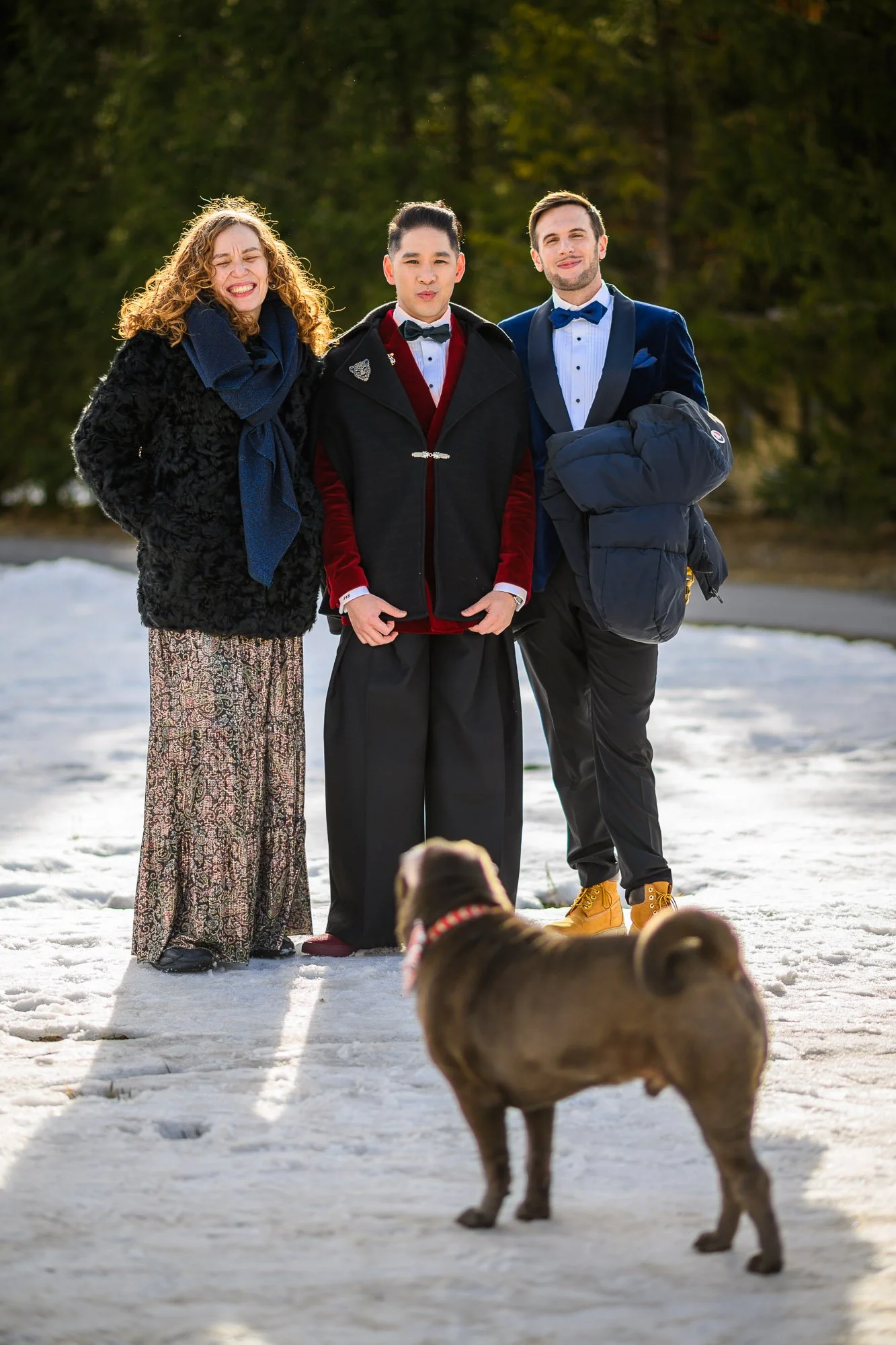 Trois personnes habillées élégamment posent en extérieur dans la neige, avec un chien brun en premier plan. Arrière-plan d'arbres verts.