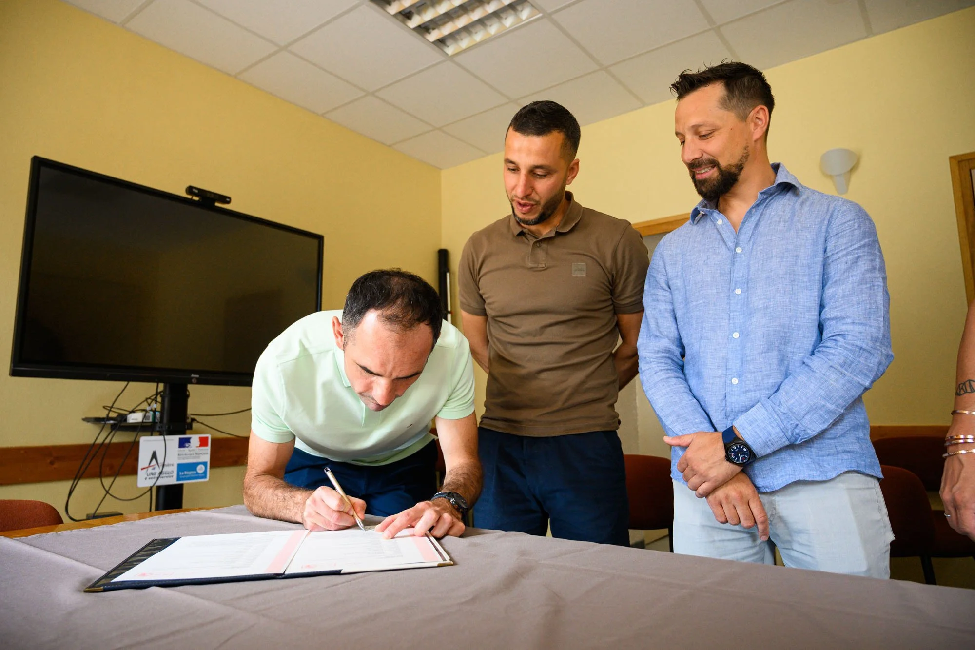 Un homme signe un document sur une table alors que deux autres hommes le regardent et l'observent dans une pièce.