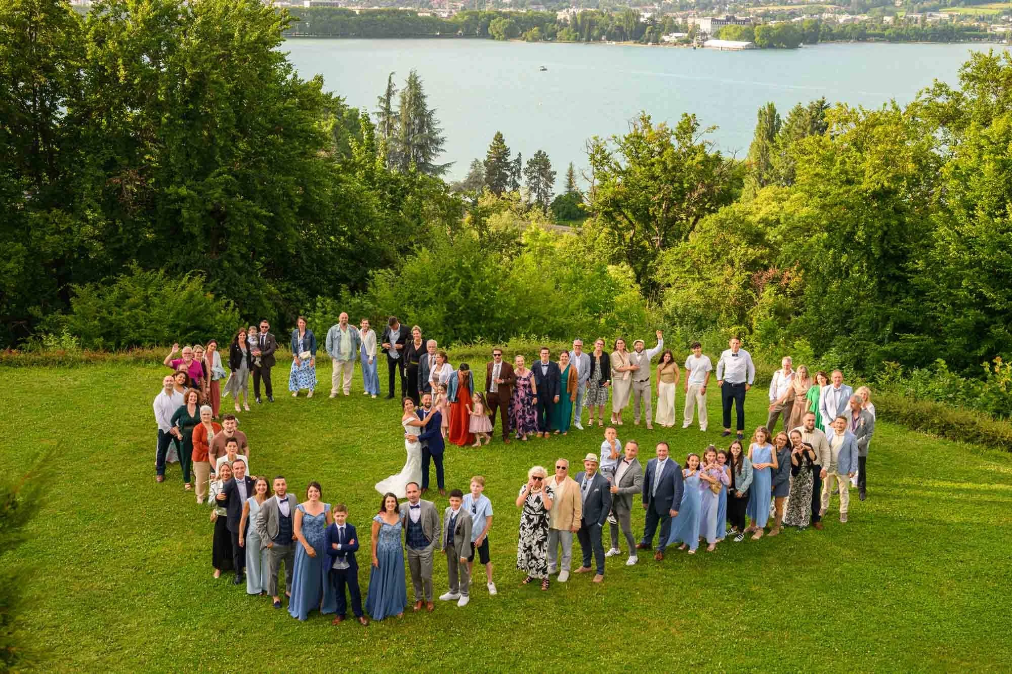 Groupe de personnes lors d’un mariage en plein air formant un cœur sur une pelouse, avec un lac et des arbres en arrière-plan.