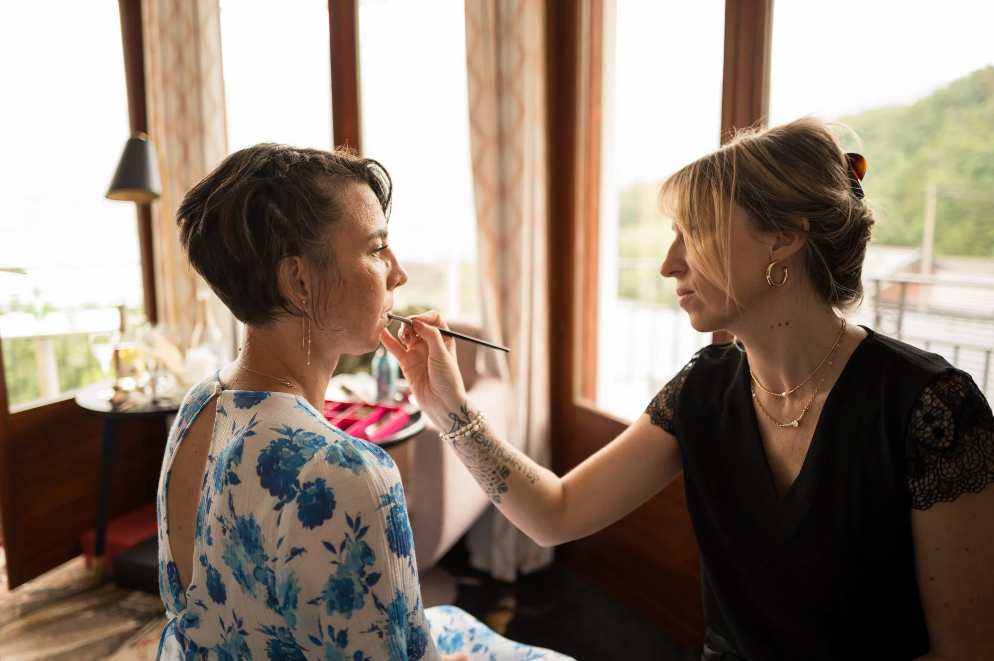Une femme avec des cheveux courts et une robe blanche à motifs bleus se fait maquiller par une autre femme aux cheveux blonds coiffés en chignon, portant un haut noir avec des détails en dentelle, dans une pièce en bois avec de grandes fenêtres laiss