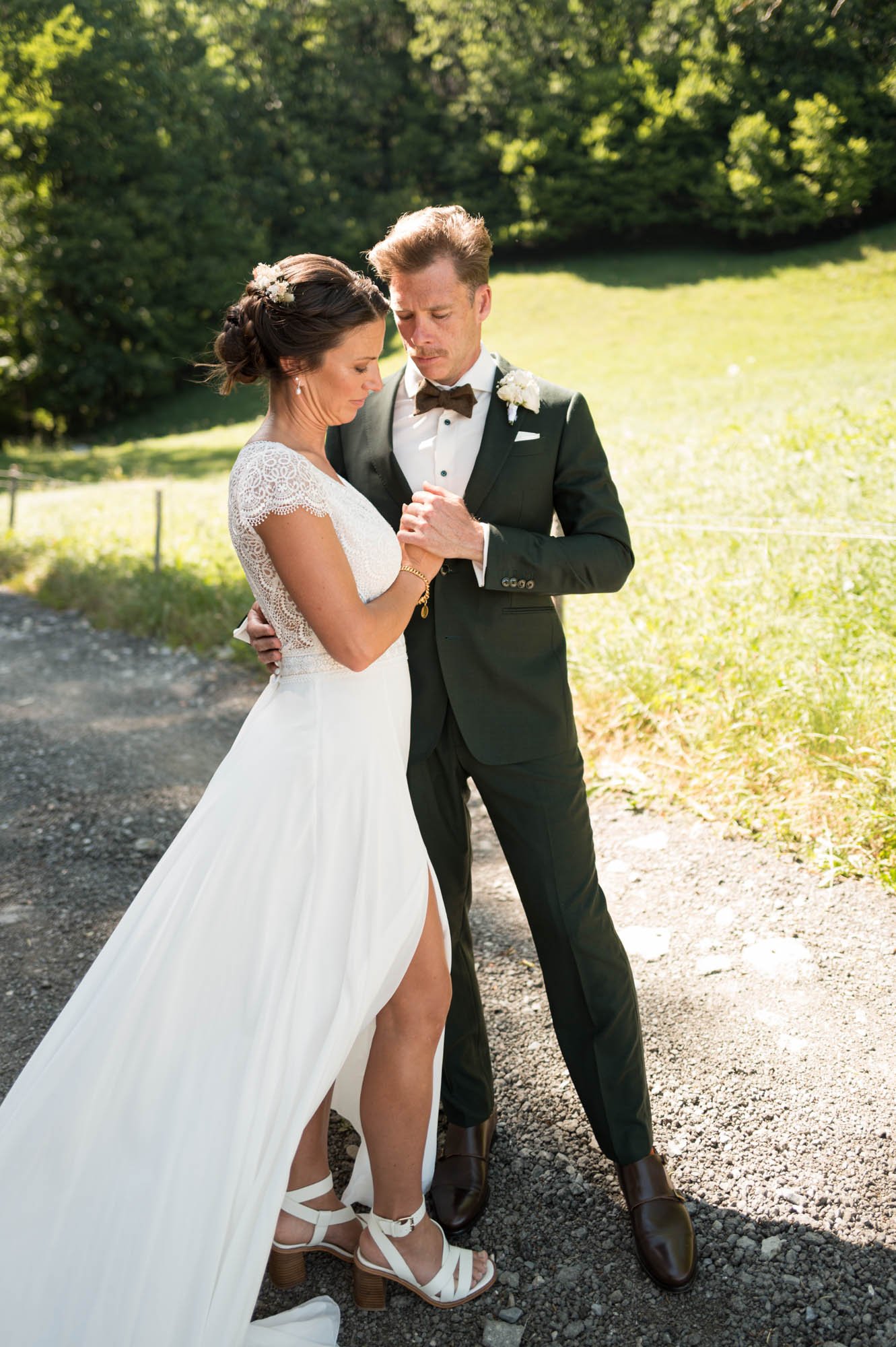 Un couple marié se tient dans un cadre naturel en plein air, la femme porte une robe de mariée blanche avec des talons blancs et le homme porte un costume noir avec une chemise blanche et un nœud papillon. Ils semblent en prière ou en méditation, ave