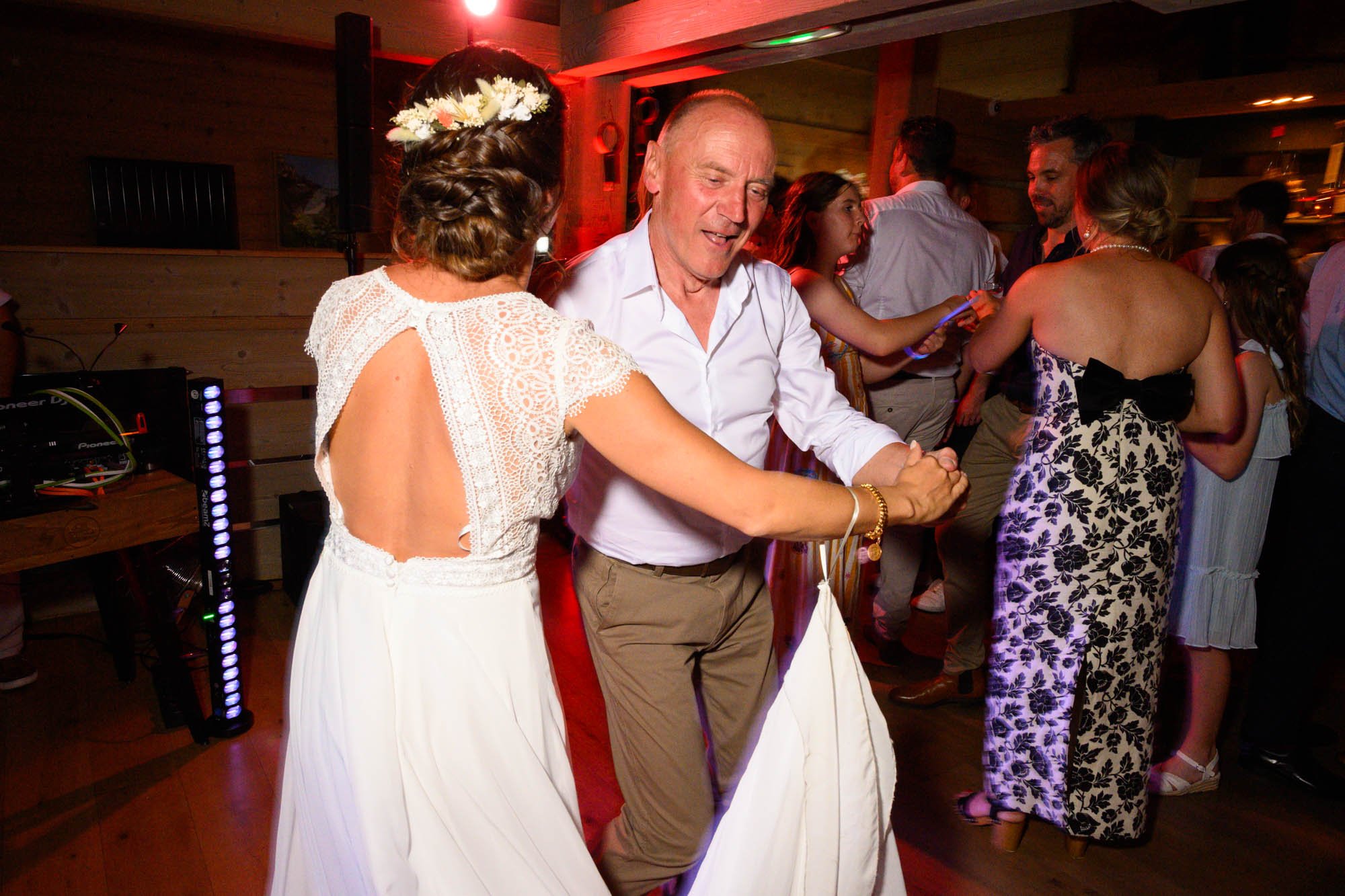 Deux personnes dansent lors d'une fête dans une ambiance chaleureuse, entourées d'autres invités. La femme porte une robe blanche avec un dos découvert, et l'homme porte une chemise blanche et un pantalon beige.