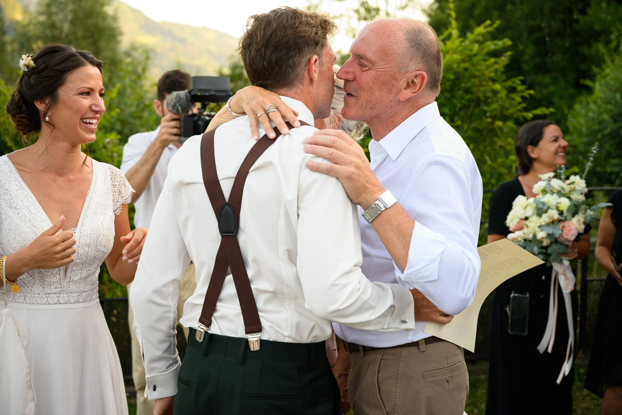 Un groupe de personnes lors d'un mariage, avec deux hommes échangeant un câlin ou un baiser, une femme souriante à gauche, une femme tenant un bouquet en arrière-plan à droite.
