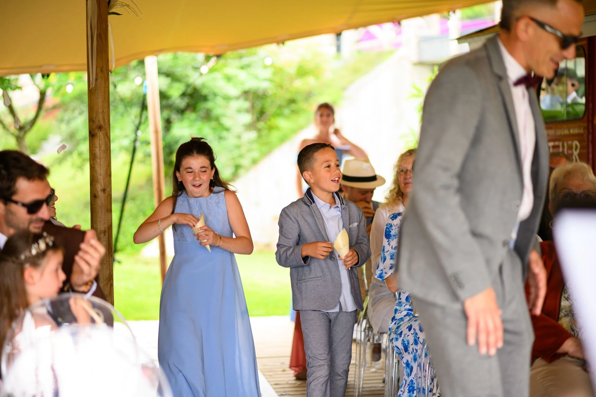 Deux enfants, un garçon et une fille, en costume et robe, souriants, tenant des cônes de glace, lors d'une cérémonie en plein air sous une tente, entourés d'adultes assis. Un homme en costume gris avec des lunettes de soleil passe devant.