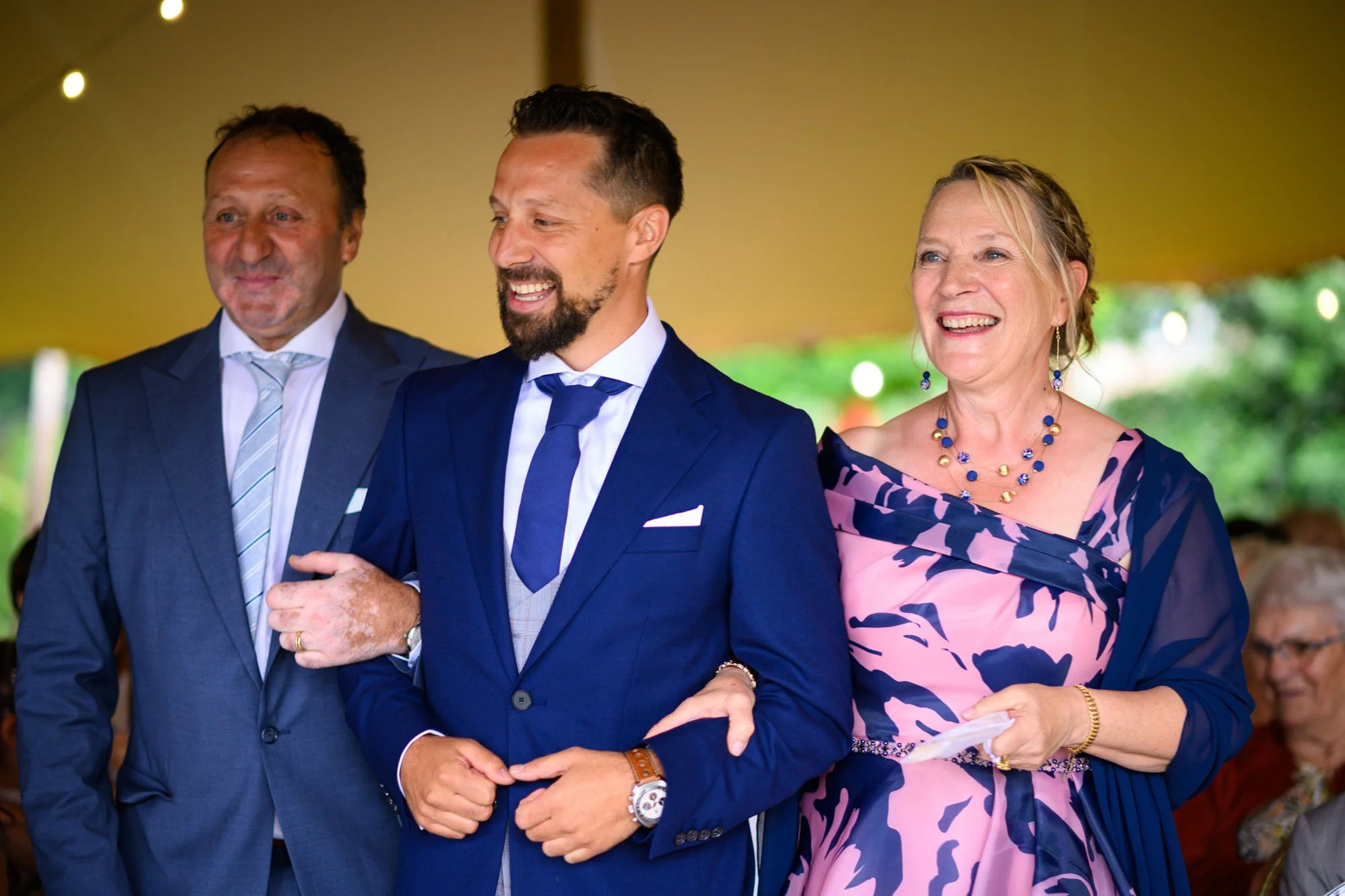 Trois personnes habillées en costume ou robe élégante, debout côte à côte, souriant lors d'un événement en plein air.