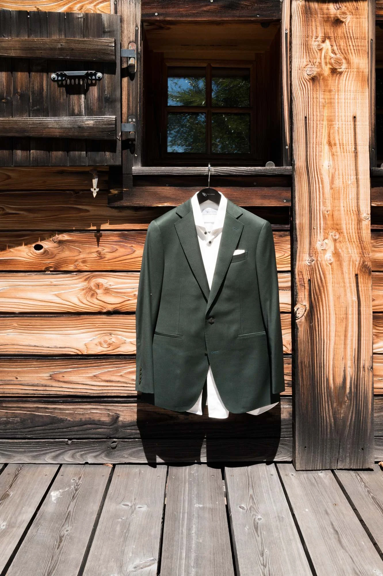 Un costume de mariage gris suspendu devant une maison en bois, avec une fenêtre et une porte en bois à l'arrière.