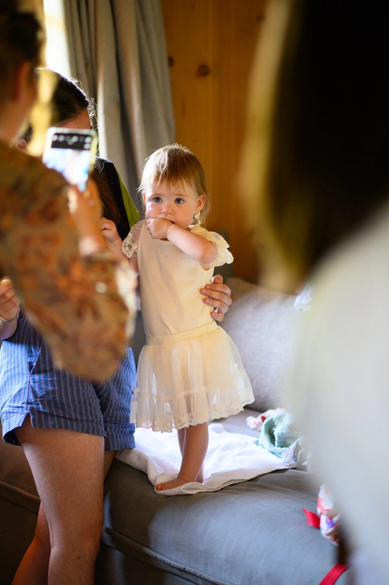 Une petite fille en robe jaune est debout sur un canapé, tenant une main d'un adulte. Elle regarde fixement l'appareil photo ou la personne en face d'elle, avec le doigt dans la bouche. Il y a d'autres adultes autour, certains prennent des photos.