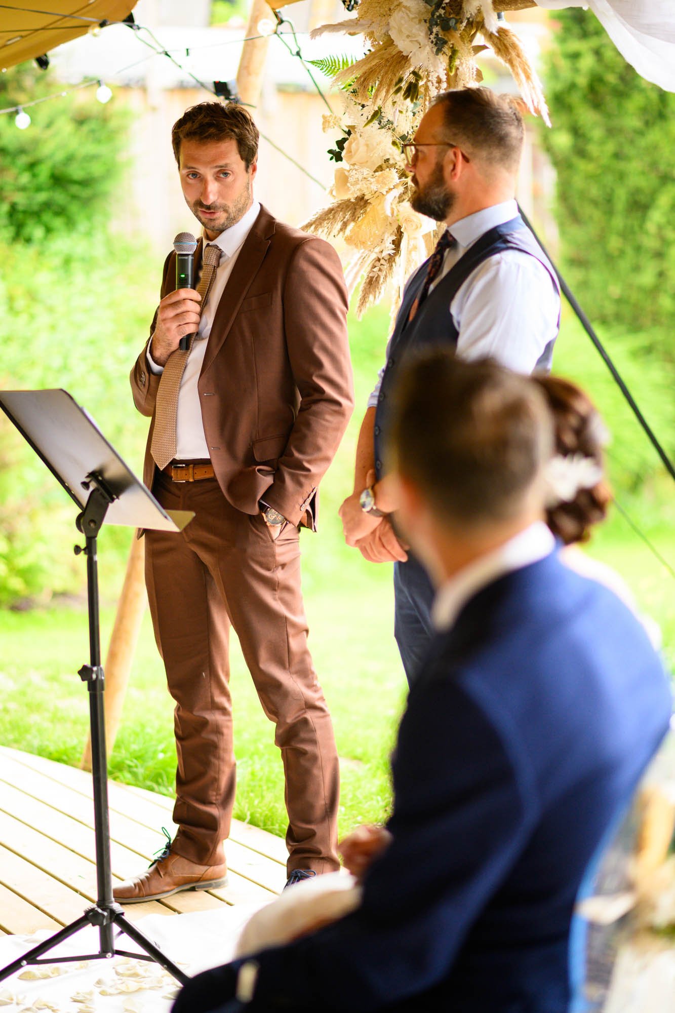 Un homme en costume marron parle dans un micro lors d'une cérémonie extérieure, avec d'autres personnes assises en face, dans un cadre verdoyant.