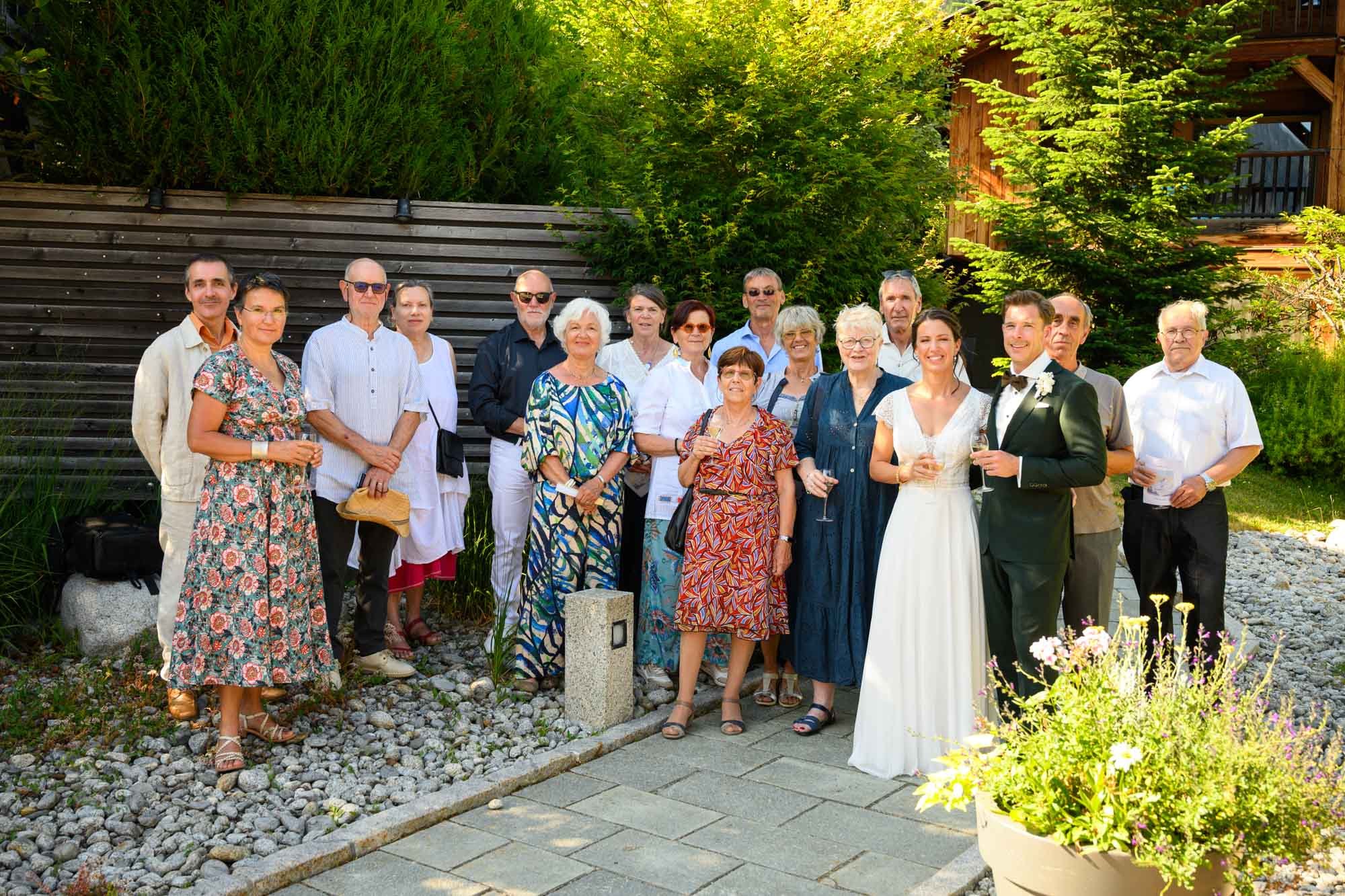 Groupe de personnes lors d'une célébration en extérieur, dont un couple en tenue de mariage. L'arrière-plan comprend des arbres verts et un bâtiment en bois.