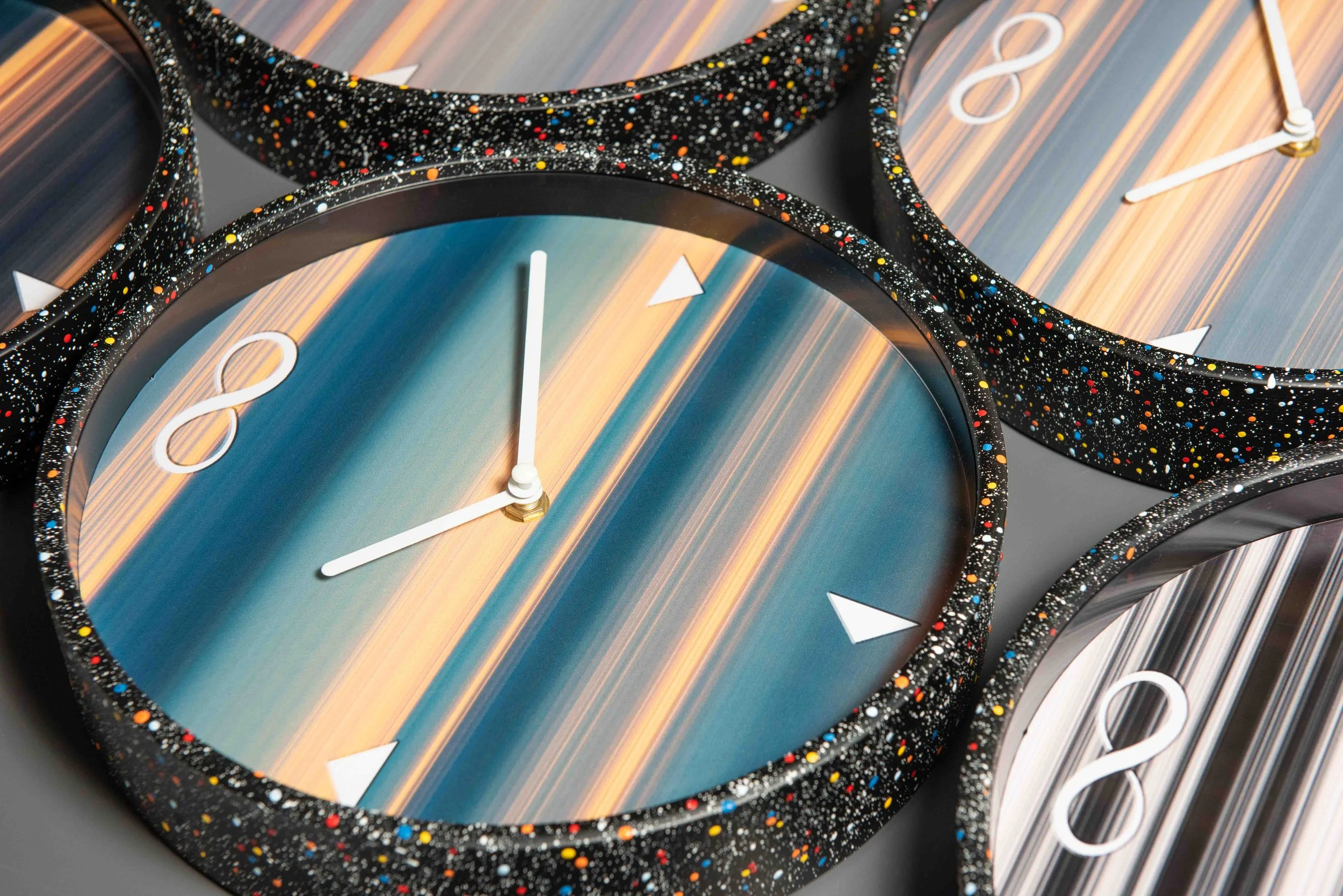 Close-up view of several circular Now Clock artworks arranged together, showing vertical bands of light, white clock hands, infinity symbols, and black bezels painted with multicolored star patterns.
