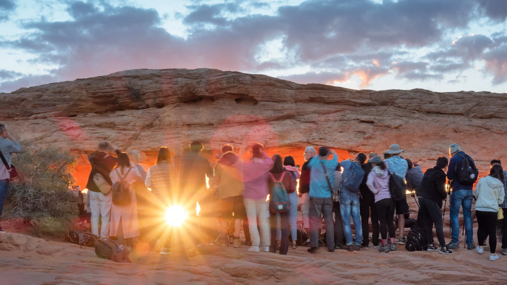 A large group of people gathered outdoors at Mesa Arch for sunrise. The sun is rising in front of them, creating a bright lens flare.