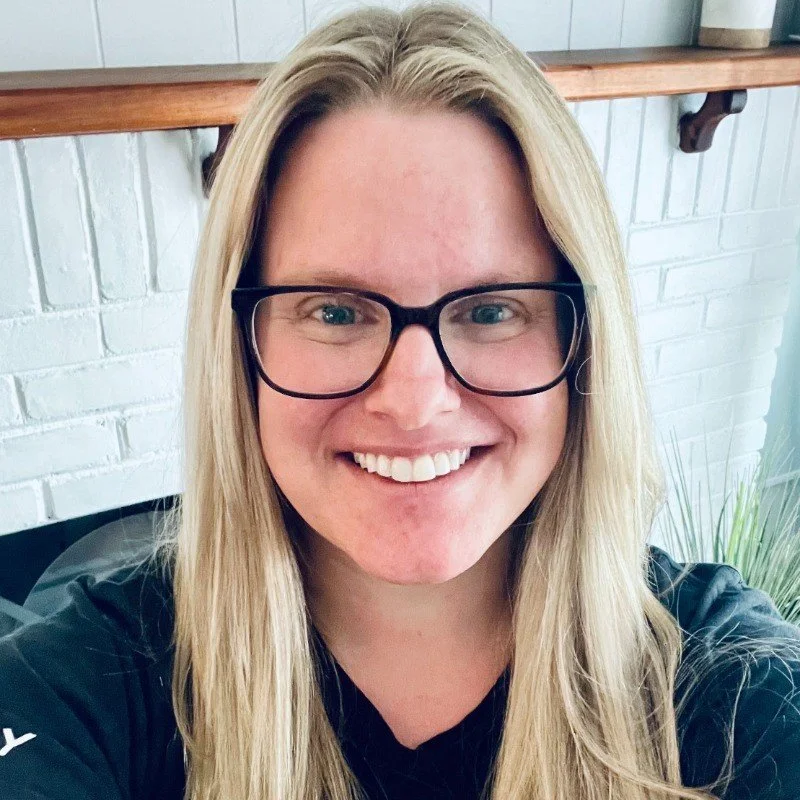 Close-up of a woman with long blonde hair, black glasses, smiling, wearing a black shirt, against a white brick background with a wooden shelf.