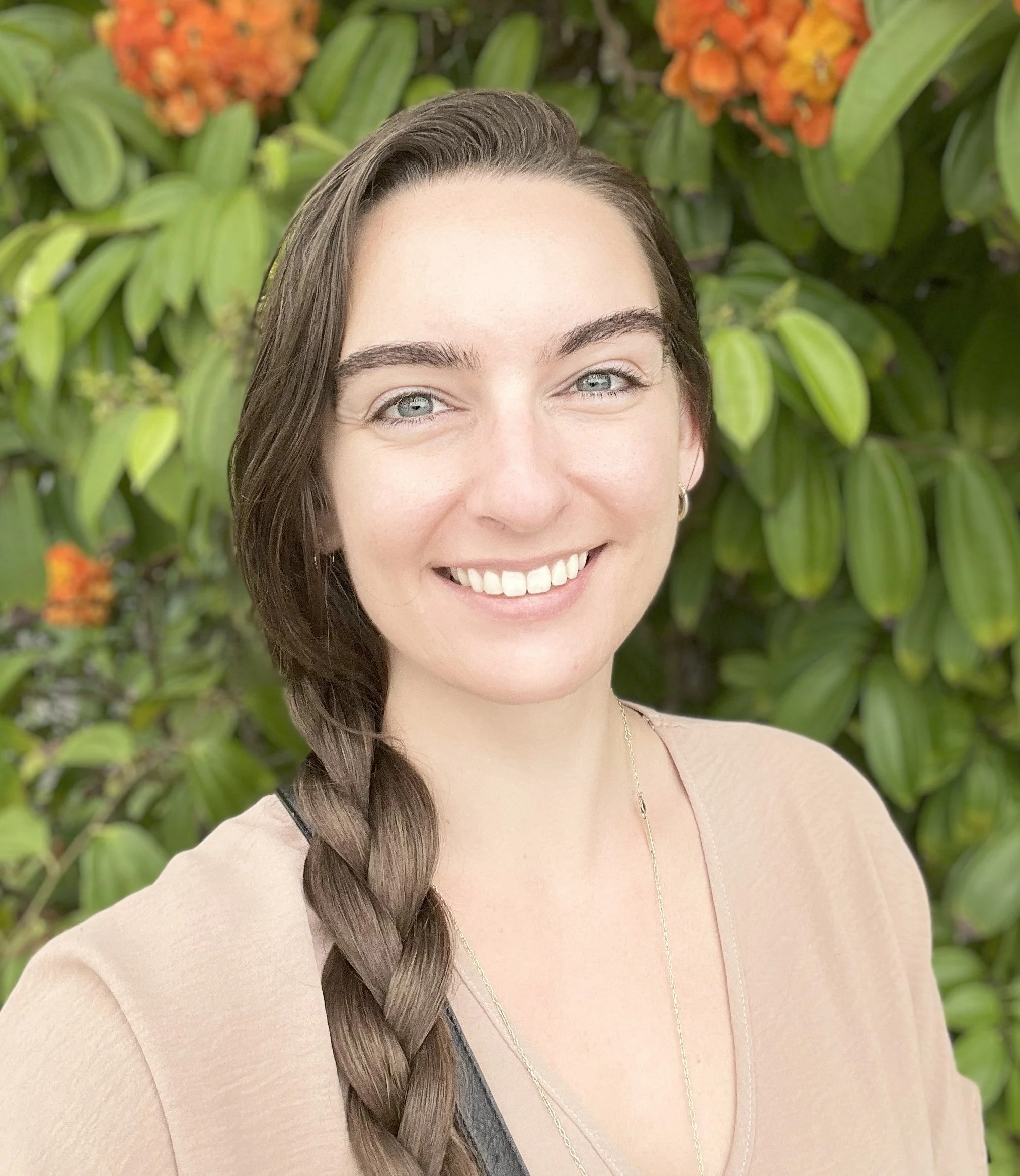 A young woman with long brown hair in a braid, smiling, standing outdoors against green leafy plants with orange flowers.