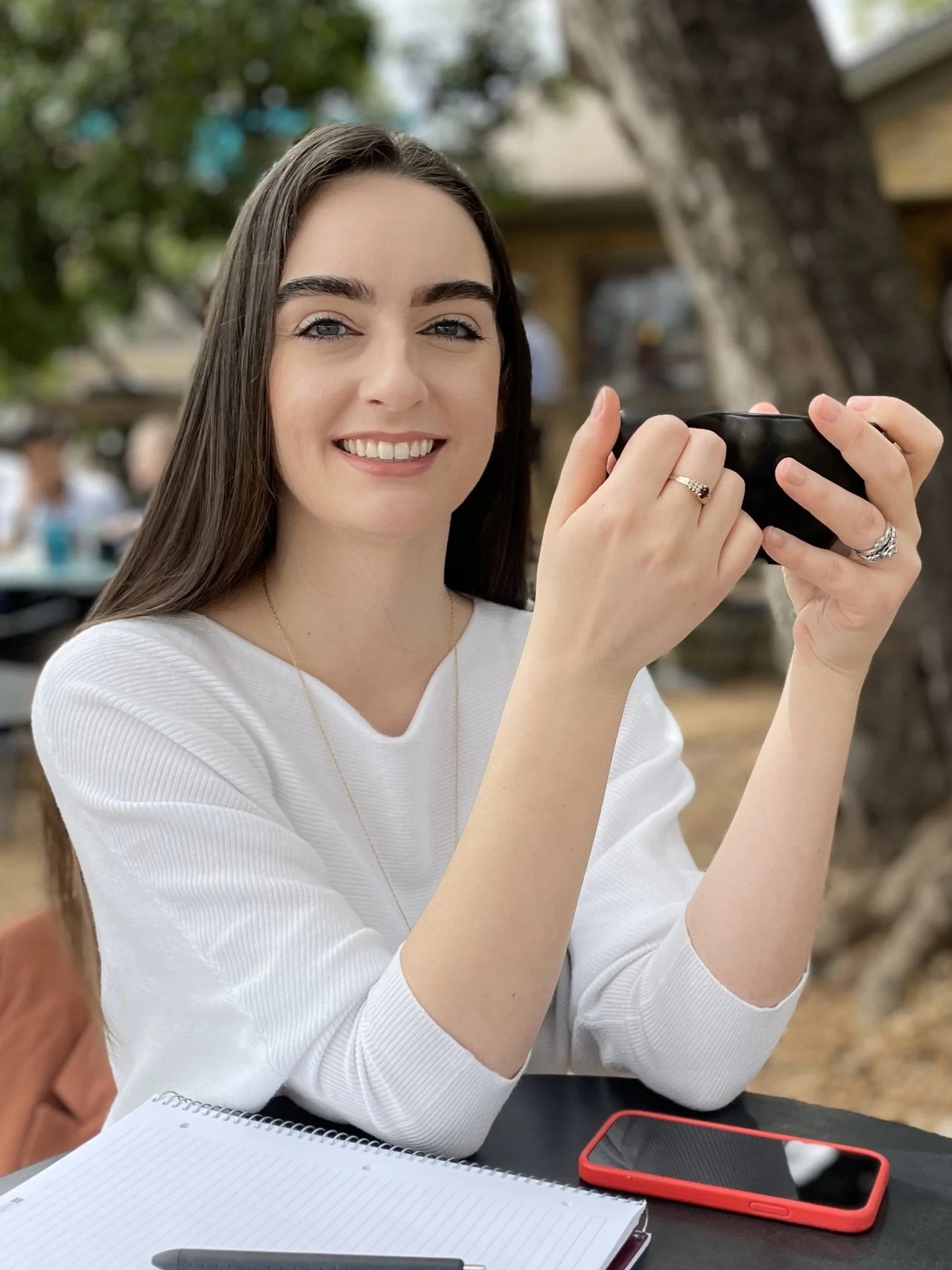 A young woman with long brown hair and blue eyes smiling while holding a black ceramic cup outdoors at a table with a notebook, pen, and red smartphone in front of her.