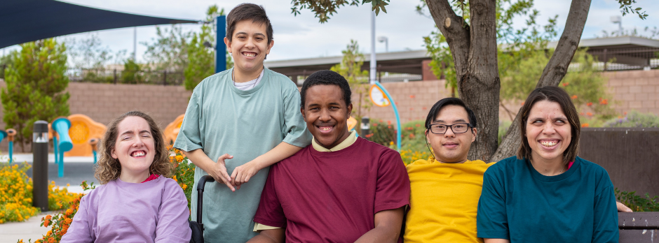 A group of five diverse young adults sitting outdoors, smiling, in a park with colorful flowers and a playground in the background.