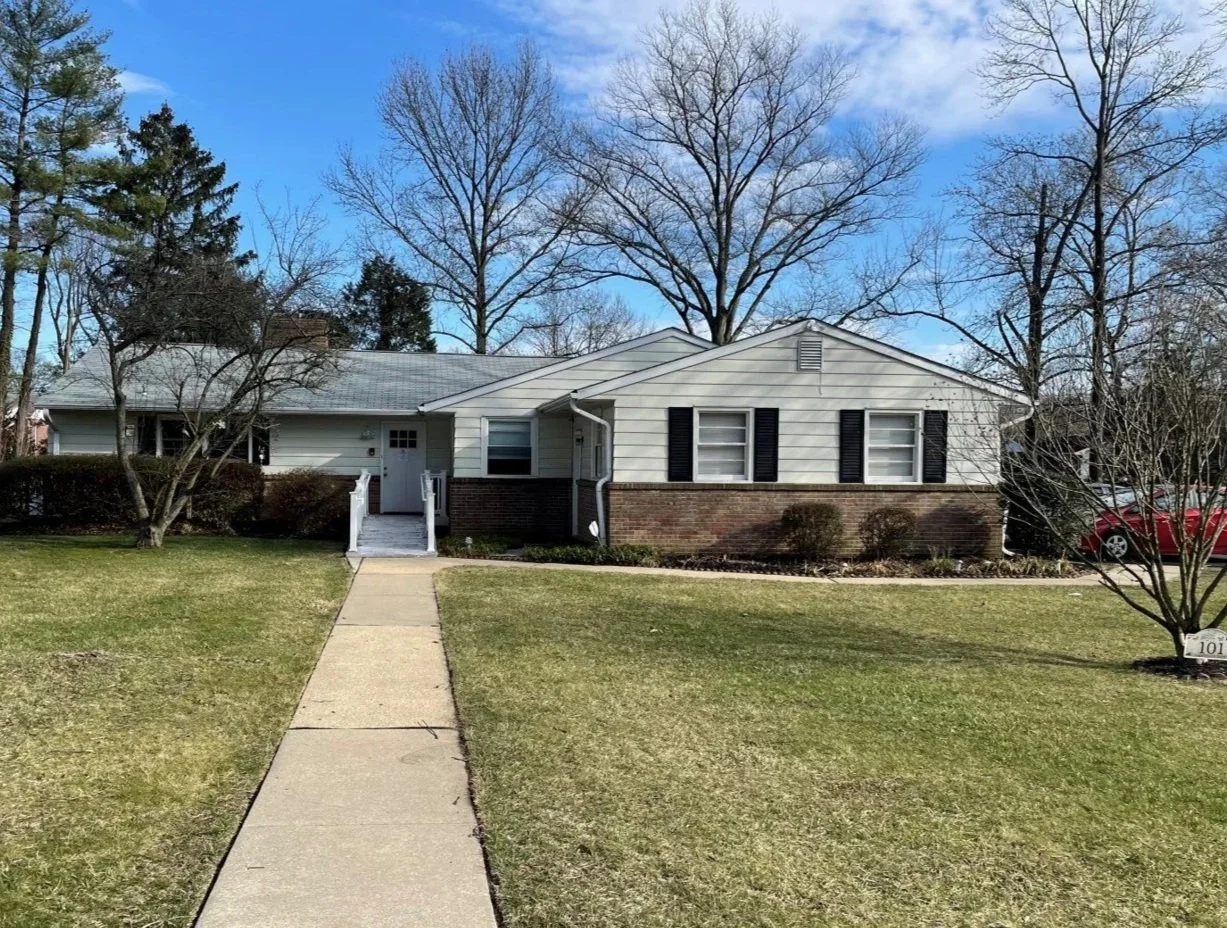 A single-story house with white siding, dark shutters, and a brick foundation, surrounded by a well-maintained lawn and trees without leaves, under a partly cloudy blue sky.