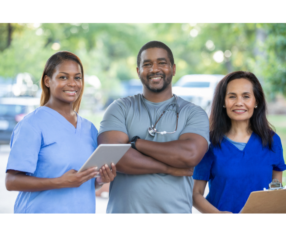 Three healthcare professionals standing outdoors, smiling, wearing scrubs, with one holding a tablet and another holding a clipboard.