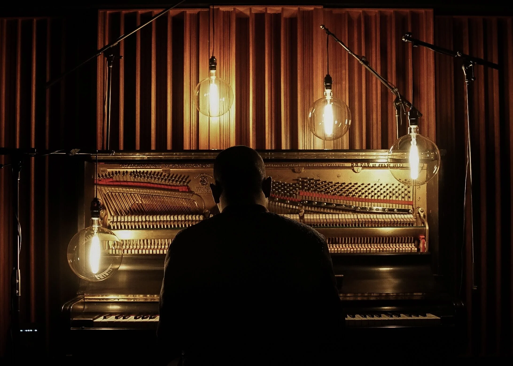 Pianist seen from behind at an upright piano beneath glowing hanging bulbs
