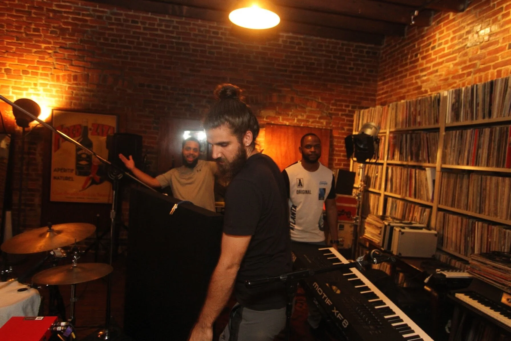 Musicians gathered around drums and keyboards in a brick-walled studio