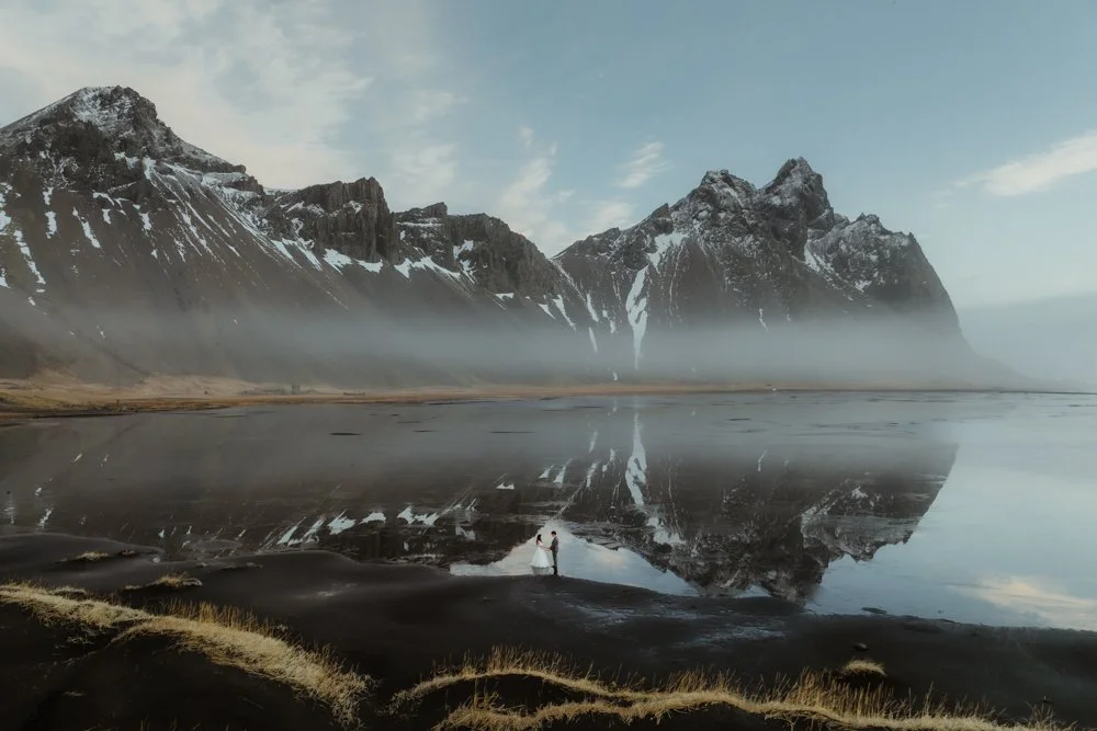 A couple holding hands near the shoreline of a reflective black sand beach, with tall, snow-capped mountains and mist in the background.
