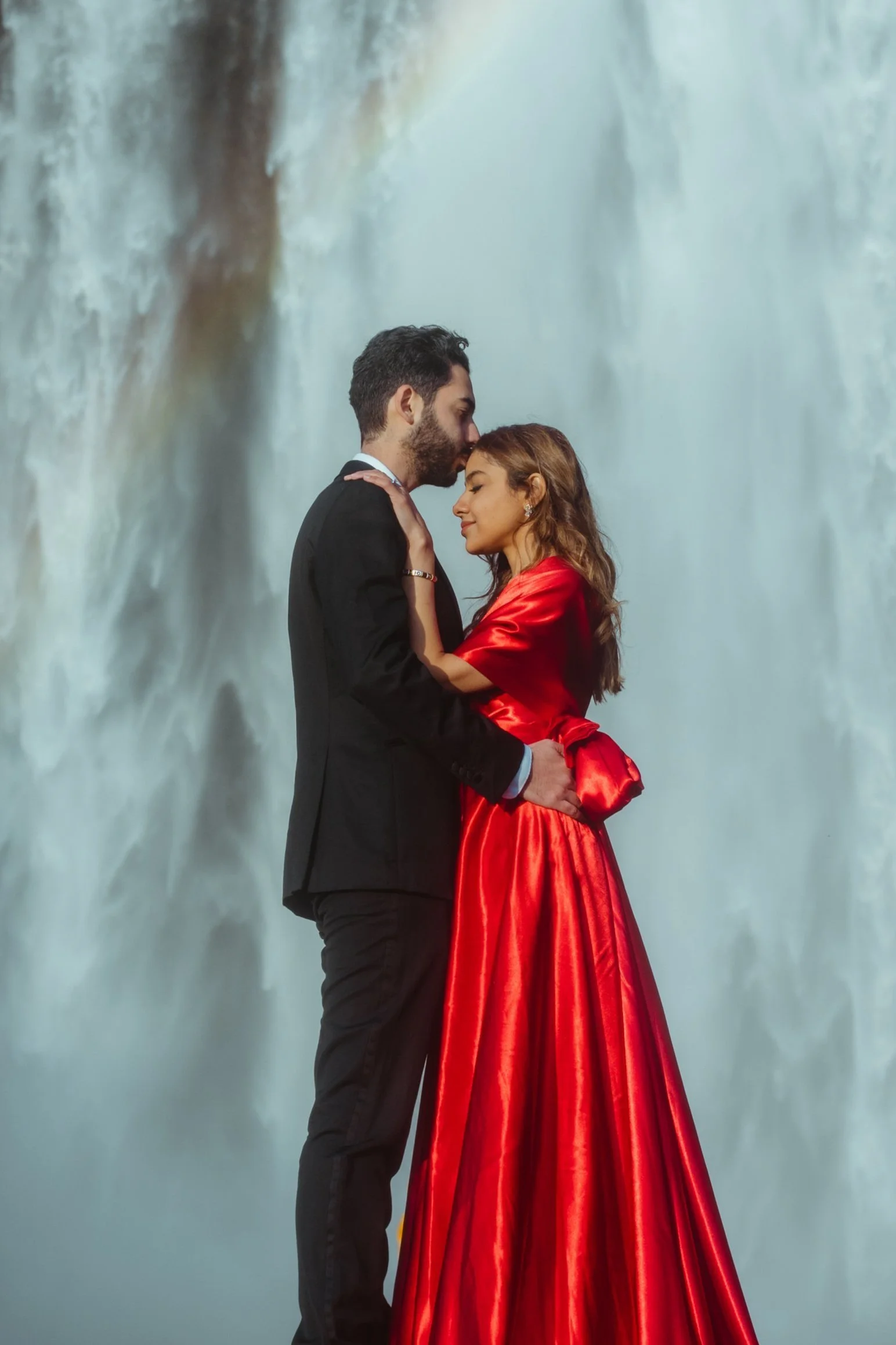 A man and woman embrace romantically in front of a waterfall backdrop; the woman wears a shiny red gown, and the man wears a black suit.