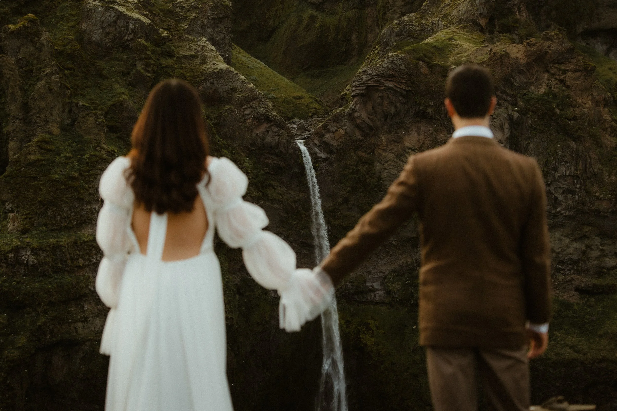 A couple holding hands, facing a waterfall in a mountainous landscape.