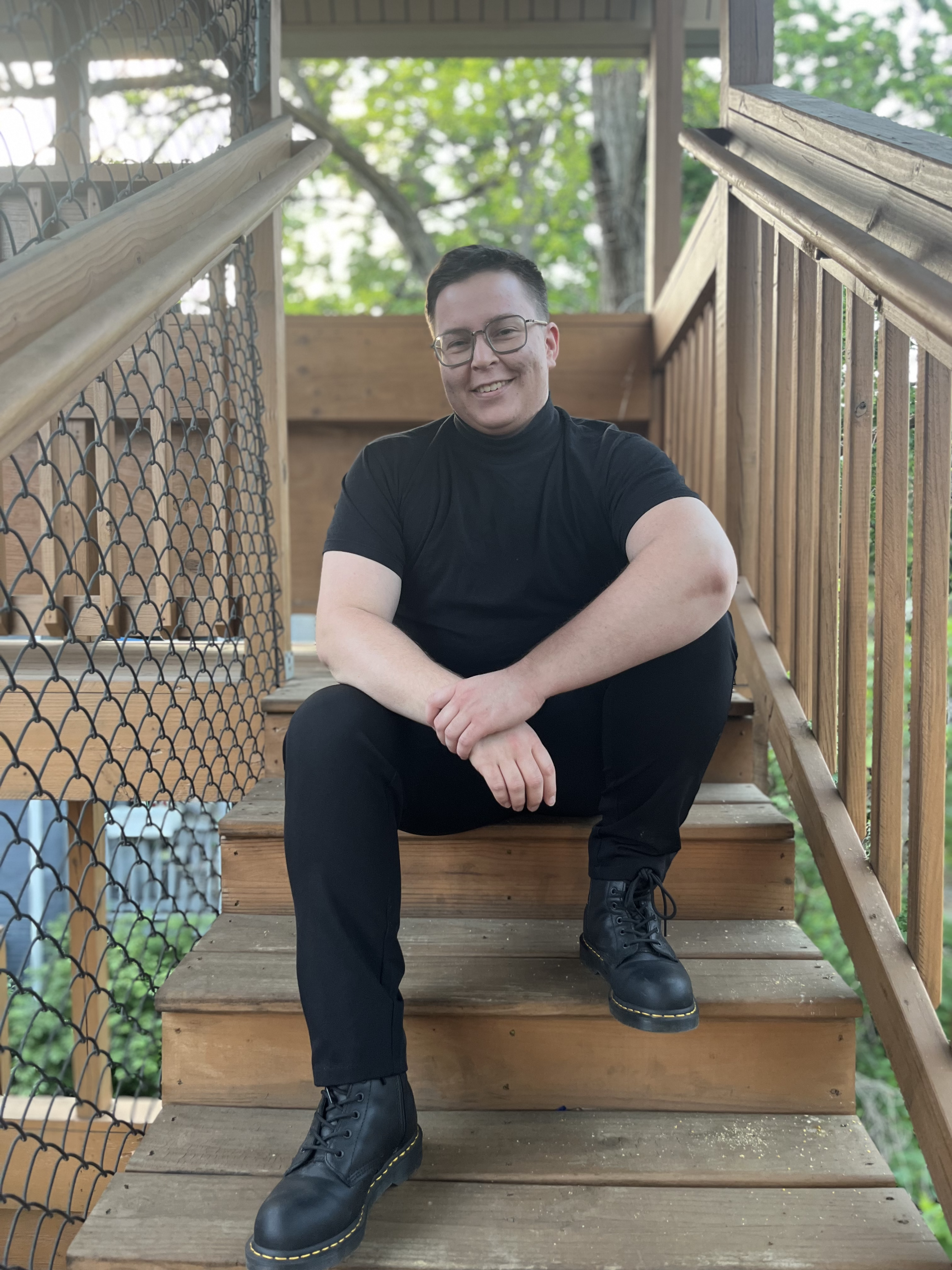 Gabriel sits on a wooden outdoor staircase. He has short dark hair and glasses. He wears a black mock-neck shirt, black pants, and black boots, with his arms resting on his knees. He's smiling warmly at the camera. Green foliage is in the background.
