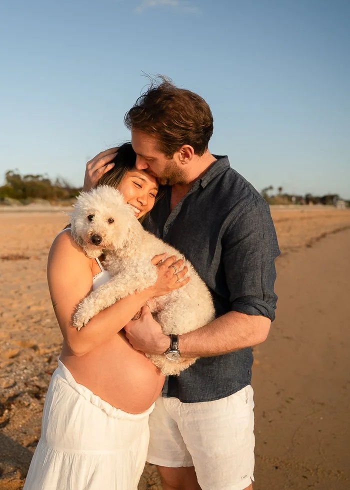 Melbourne maternity photographer photo of a pregnant woman showing her bare belly hugging her white poodle dog and her partner at the beach in golden hour