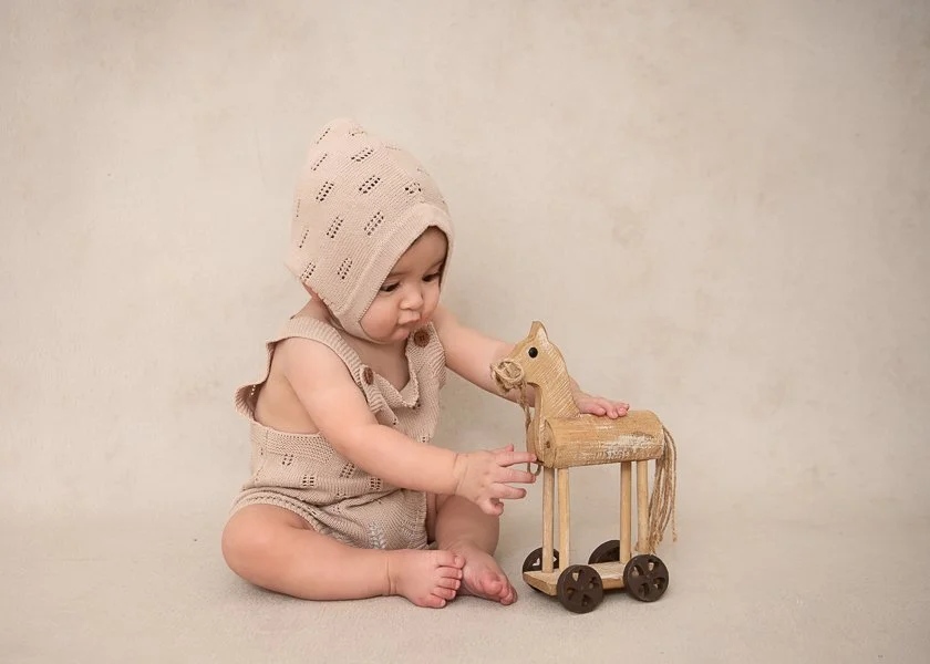 Melbourne baby photographer image of a 7 month old baby boy wear ing knitted overalls and a bonnet playing with a wooden horse in a Brighton studio setting