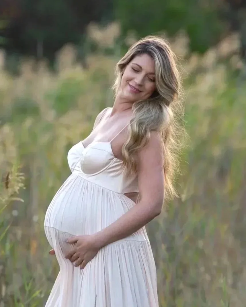 A pregnant woman with long, wavy blonde hair in a white dress standing in a natural outdoor setting, gently holding her belly and smiling with her eyes closed.