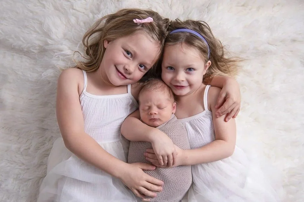 Two young girls with long hair and headbands hugging a newborn baby on a fluffy white blanket.